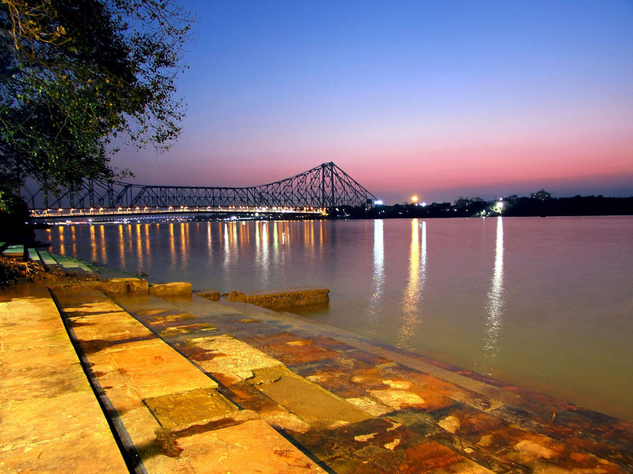 Kolkata 桌布 壁紙 — 一座雄偉的鋼桁架橋在黃昏時分用溫暖的燈光照亮平靜的河流，其倒影在水面上閃爍。
