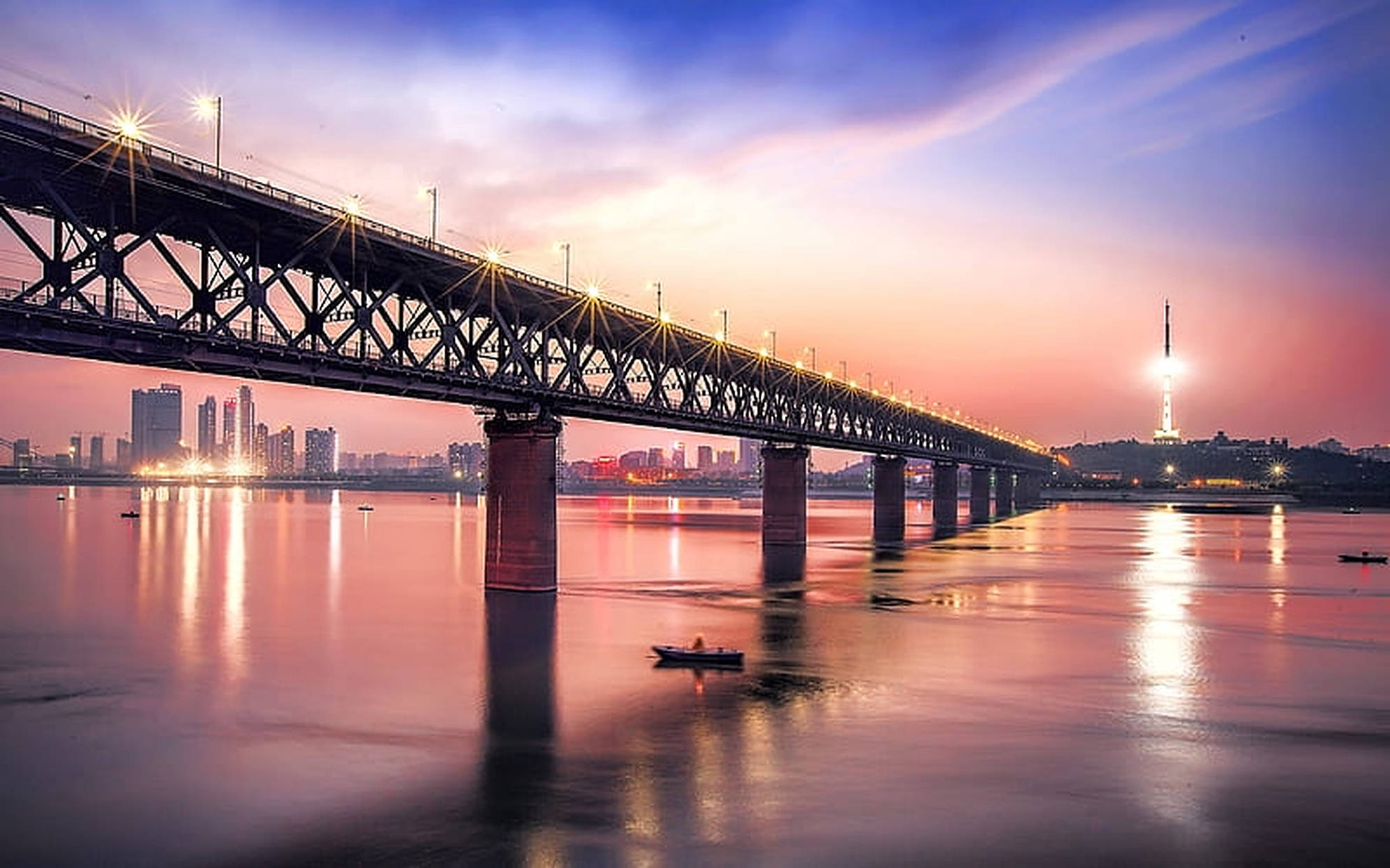 Wuhan 桌布 壁紙 — 一座雄偉的鋼橋在日落時分橫跨平靜的河流，城市燈光倒映在水面上，一艘孤獨的小船在下方漂流。