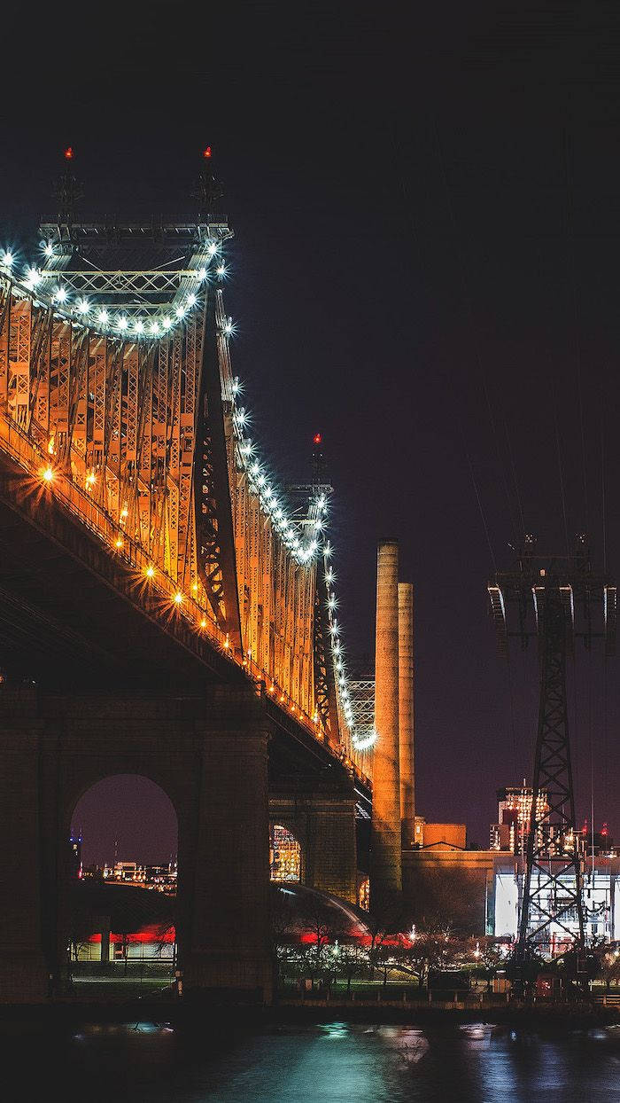 New York 桌布 壁紙 — 一座雄偉的鋼橋在漆黑的夜空下閃耀著溫暖的金色光芒，其倒影在下方平靜的水面上閃爍。