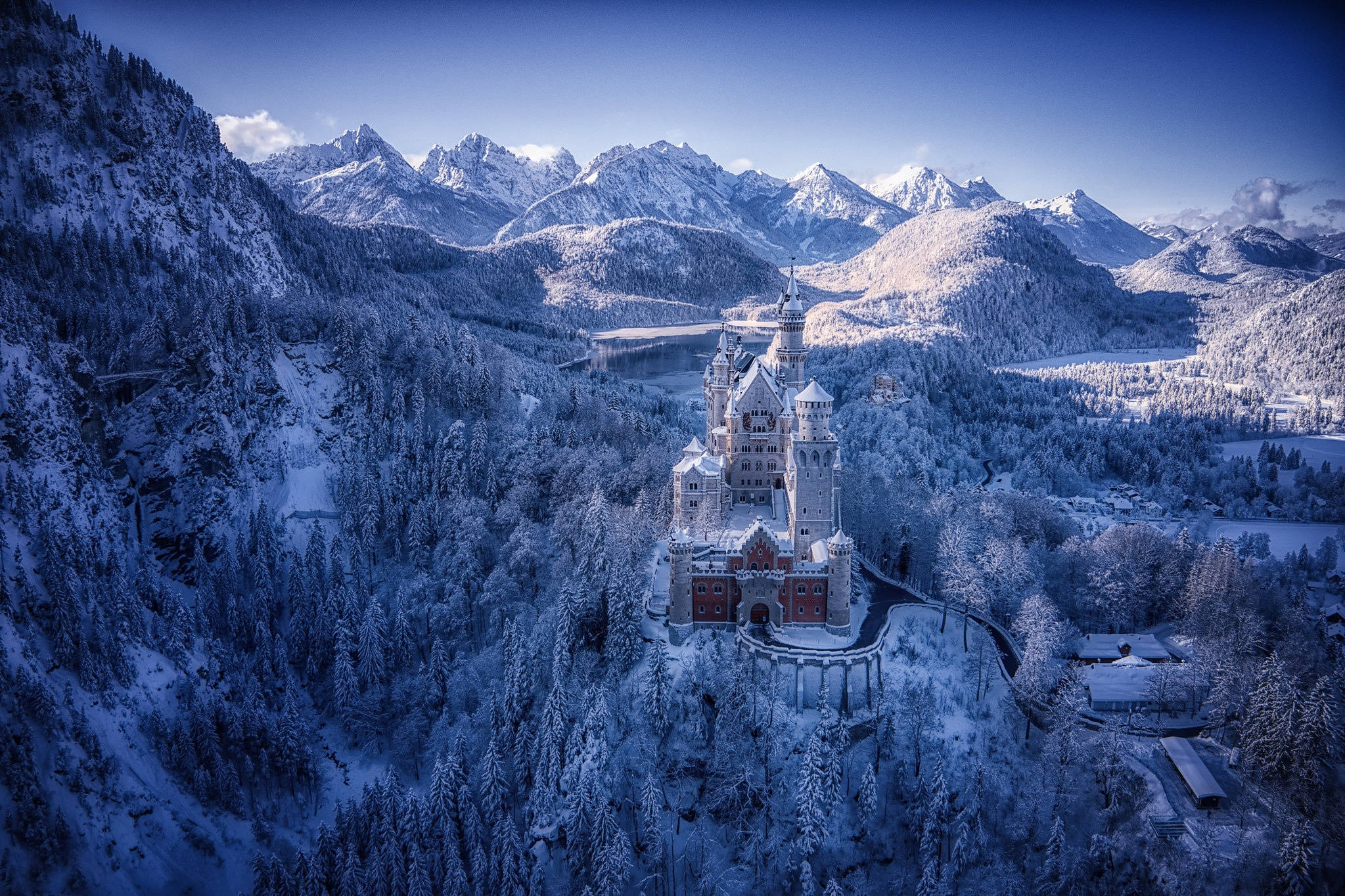 Neuschwanstein Castle 桌布 壁紙 — 一座雄偉的雪城堡坐落在山丘上，四周環繞著冰封的森林和巍峨的阿爾卑斯山峰，頭頂是清澈的藍天。