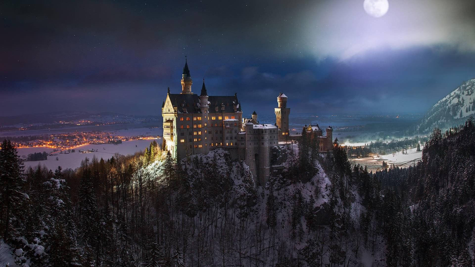 Neuschwanstein Castle 桌布 壁紙 — 一座雄偉的雪城堡坐落在山丘上，在滿月和星空下溫暖地發光。