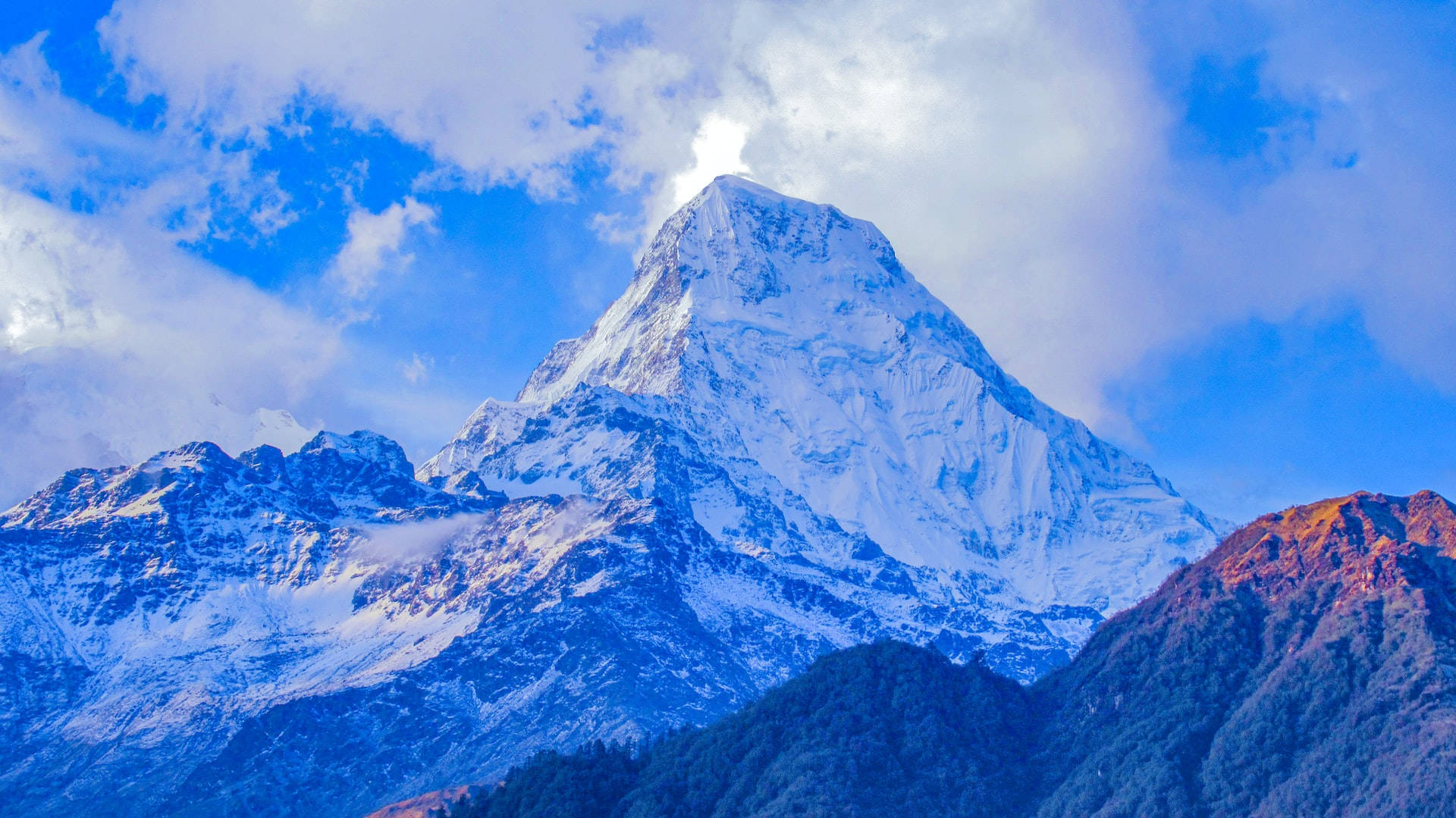 Mountain Landscape 桌布 壁紙 — 一座雄偉的雪山山峰刺破充滿活力的藍天，蓬鬆的白雲和金色的陽光突出其崎嶇的山坡。