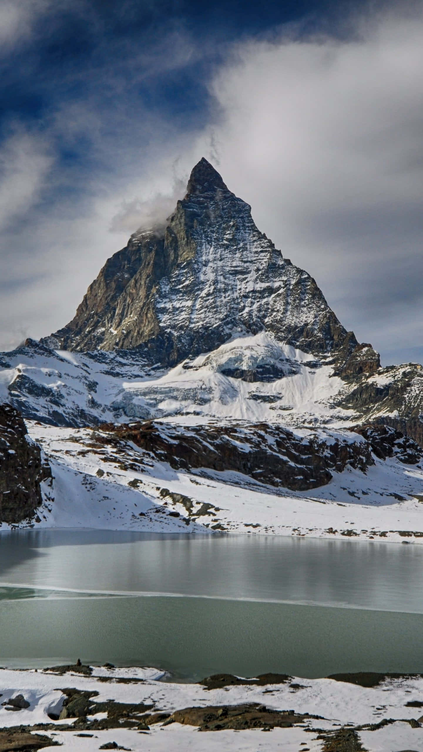 Lake 桌布 壁紙 — 一座雄偉的雪山山峰在戲劇性的多雲天空下，陡峭地聳立在寧靜的高山湖泊之上。