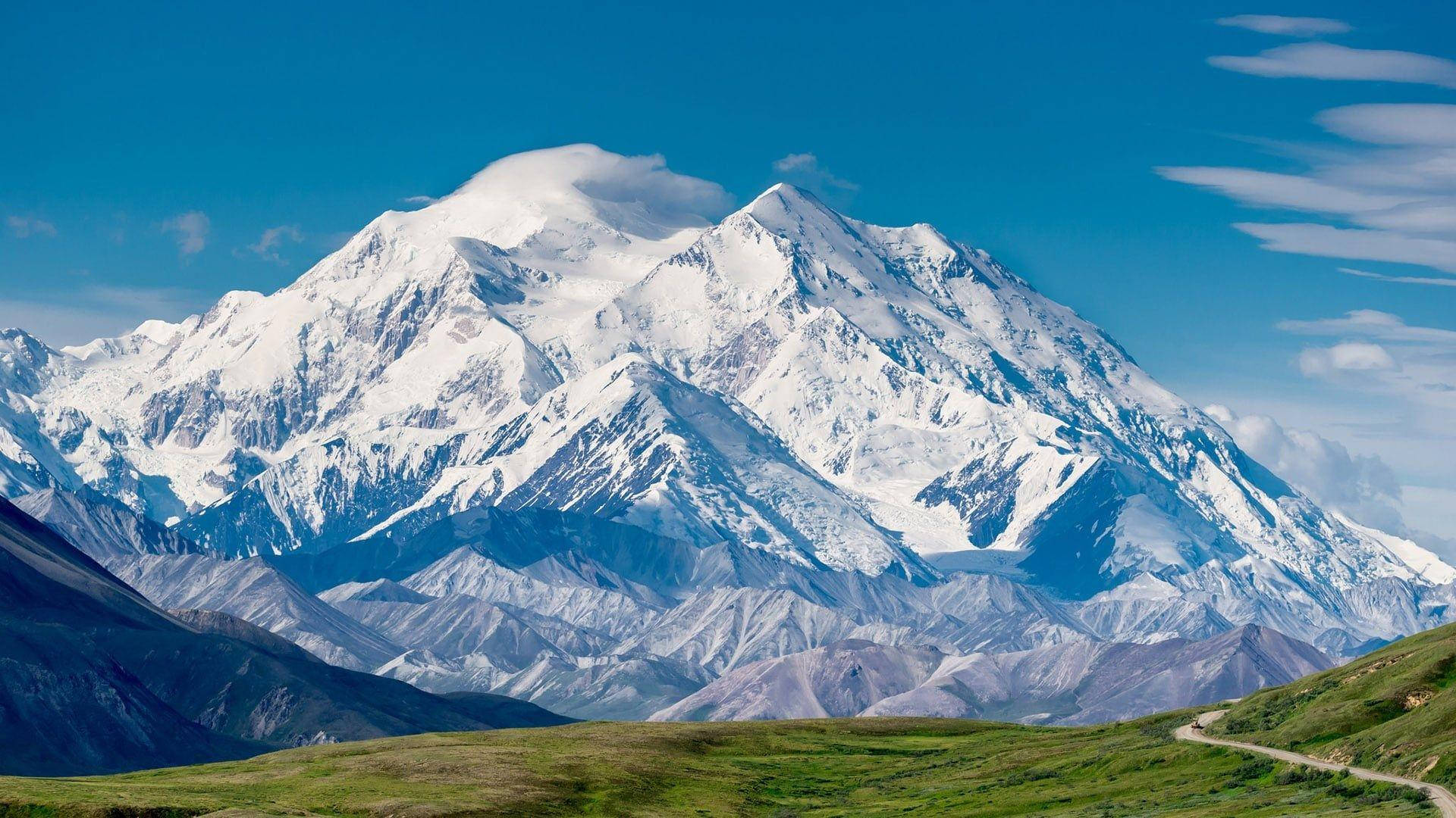 山景 桌布 壁紙 — 一座雄偉的雪山山峰在晴朗的藍天和縷縷白雲映襯下，聳立在綠色山谷和蜿蜒道路之上。