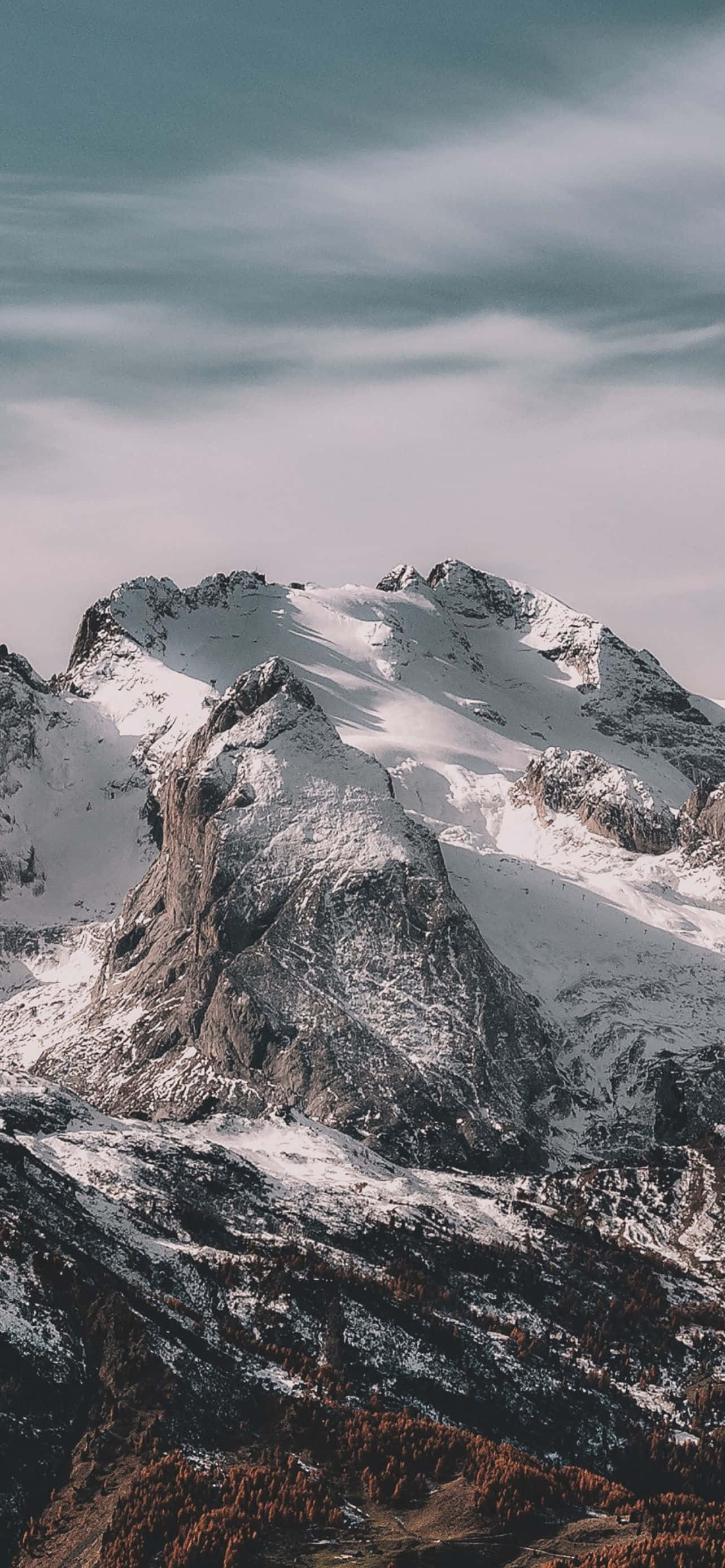 Mountain iPhone 桌布 壁紙 — 一座雄偉的雪山山峰在柔和的多雲天空和下方的秋樹襯托下巍然聳立。
