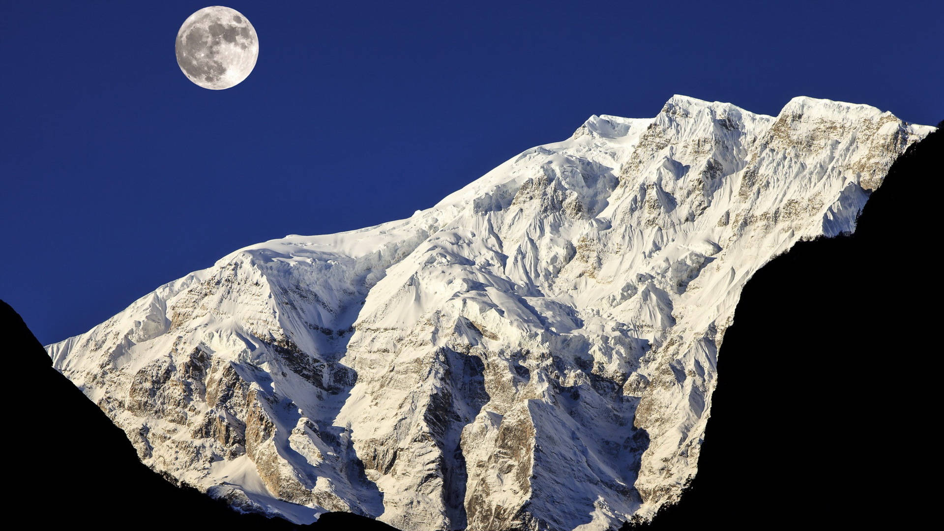 Nepal 桌布 壁紙 — 一座雄偉的雪山山峰在深藍色的天空下升起，一輪滿月在上方閃耀。