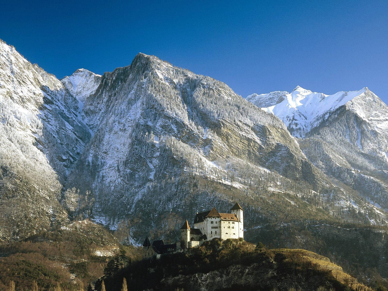 Liechtenstein 桌布 壁紙 — 一座雄偉的雪山山脈聳立在一個歷史悠久的城堡之上，城堡坐落在山坡上，背景是清澈的藍天。