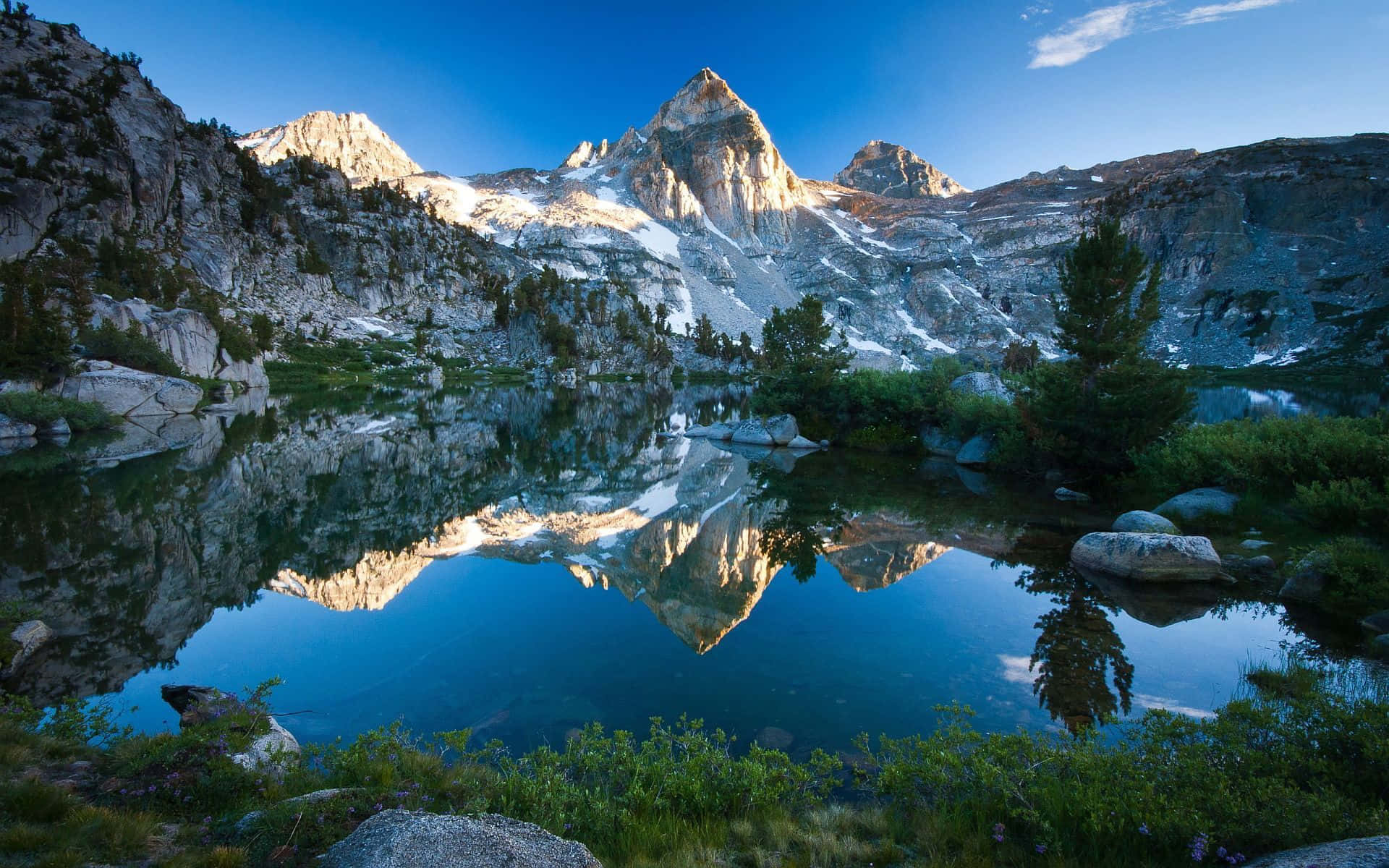 山景 桌布 壁紙 — 一座雄偉的雪山峰在寧靜的高山湖泊中完美倒映，周圍環繞著綠植和岩石，天空湛藍清澈。