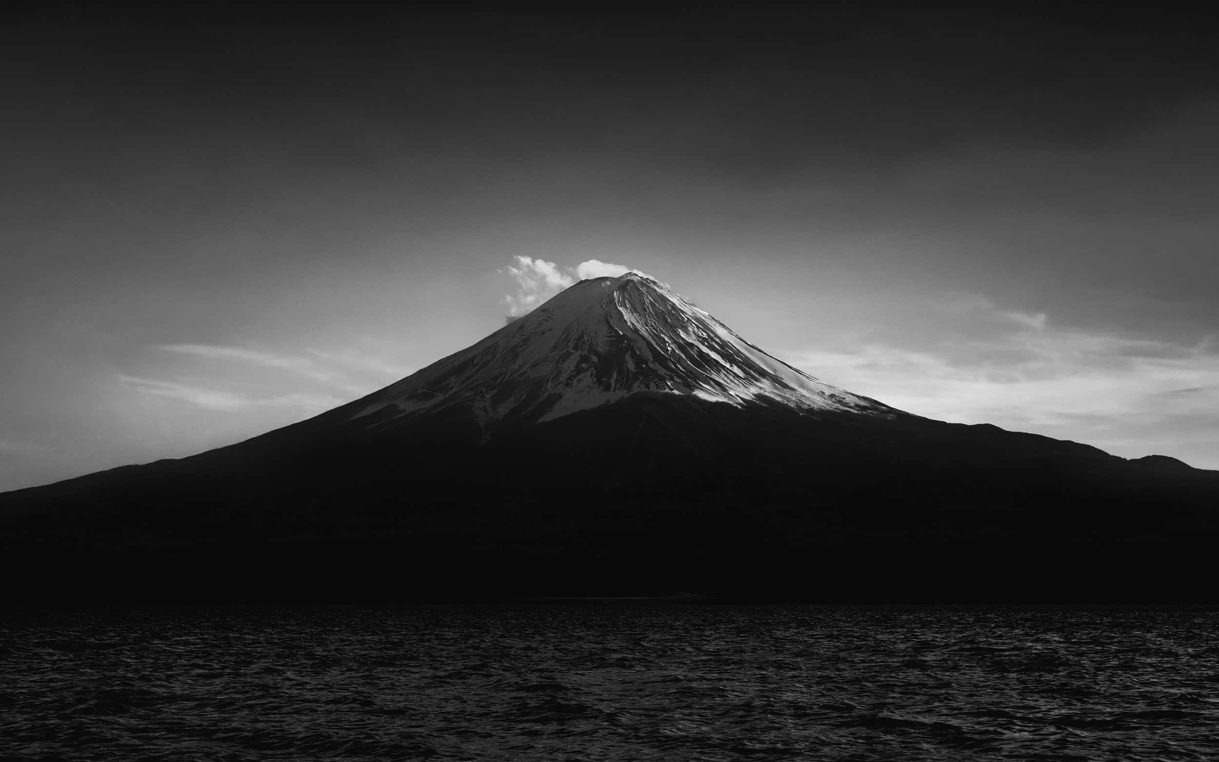 日本黑白 桌布 壁紙 — 一座雄偉的雪山從黑暗的湖泊中戲劇性地升起，在陰鬱的單色天空下。