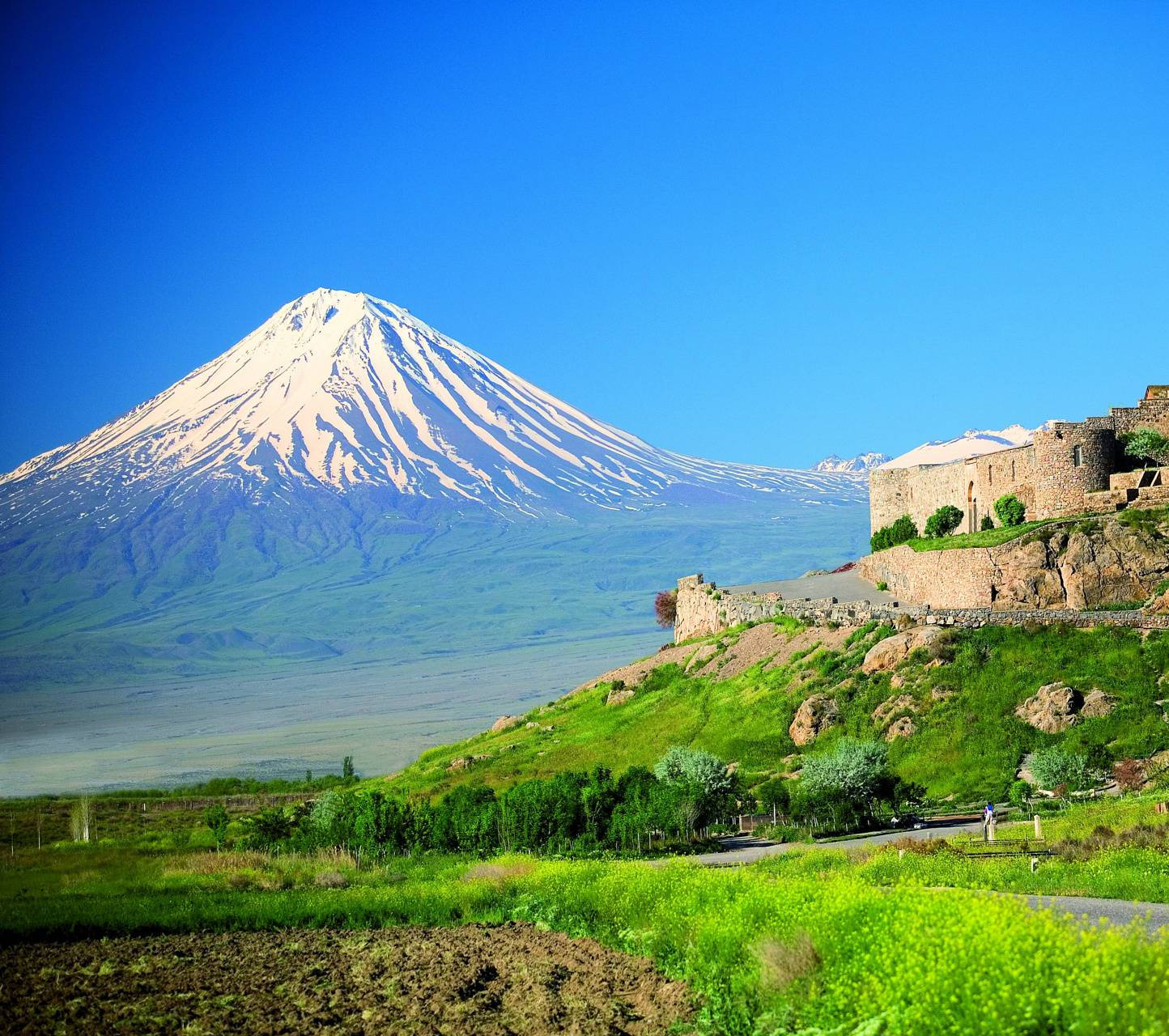 Armenia 桌布 壁紙 — 一座雄偉的雪山聳立在充滿活力的綠色山谷之上，山坡上有古老的石頭廢墟，在清澈的藍天下。