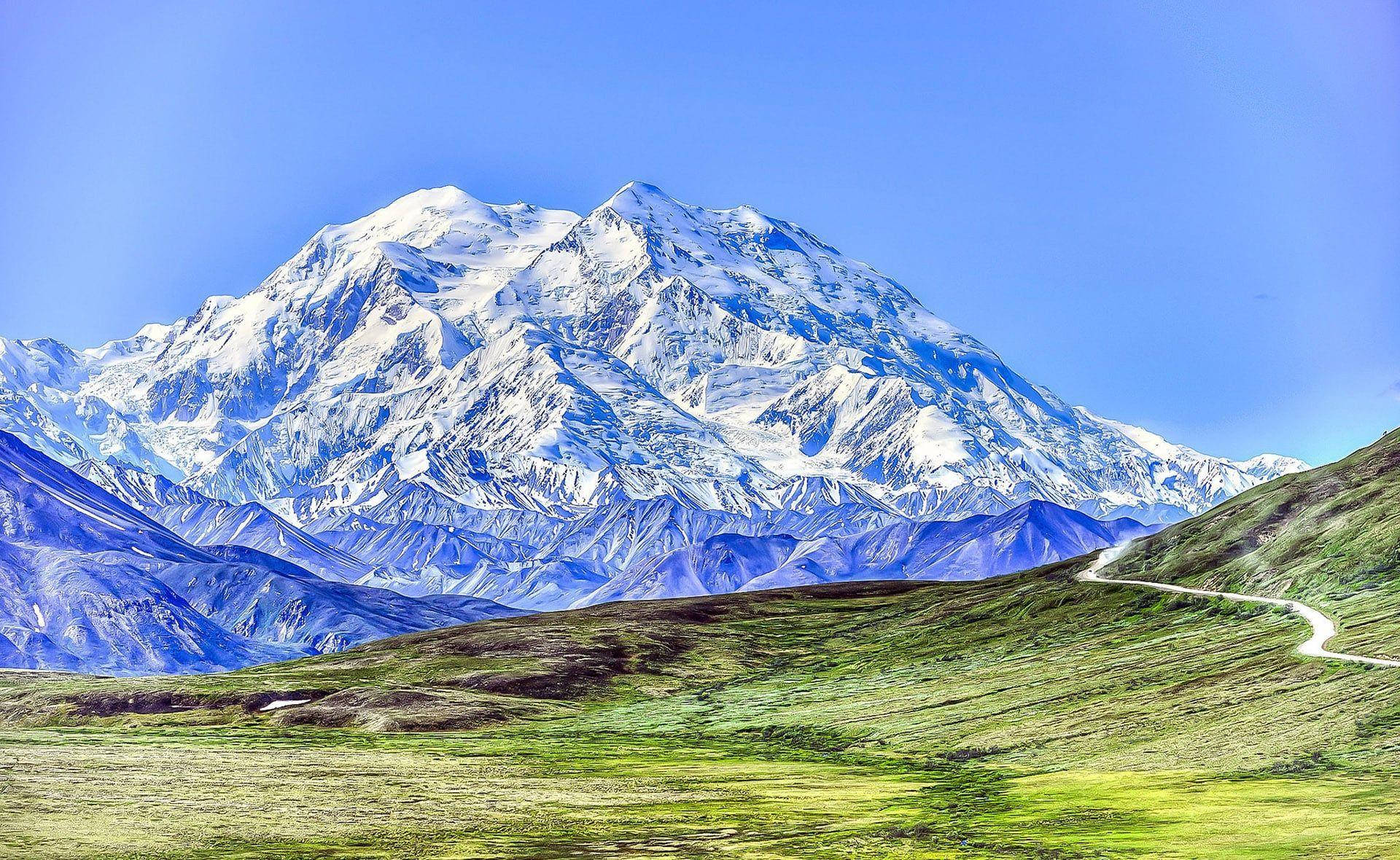 山景 桌布 壁紙 — 一座雄偉的雪山聳立在蜿蜒的道路之上，穿過充滿活力的綠色苔原，在清澈的藍天下。