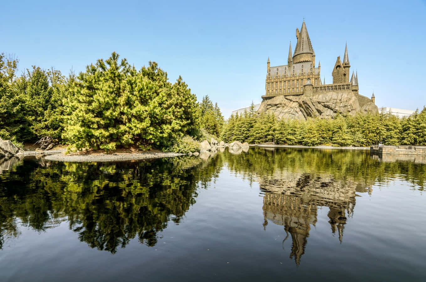 The Hogwarts Lake 桌布 壁紙 — 一座雄偉的霍格華茲城堡坐落在岩石懸崖上，完美地倒映在平靜的池塘中，池塘周圍環繞著茂密的綠樹，頭頂是清澈的藍天。