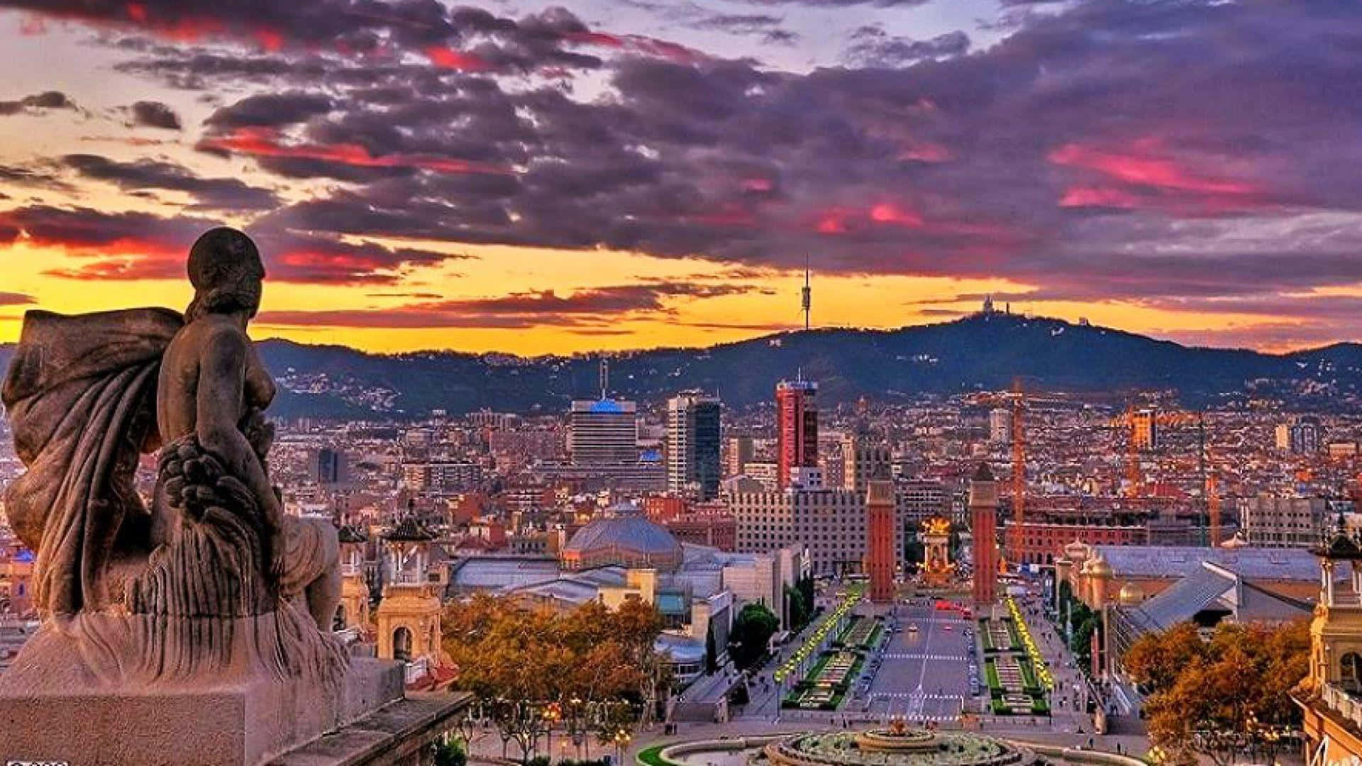 Barcelona 桌布 壁紙 — 一座雕像俯瞰巴塞隆納充滿活力的日落天際線，伴隨著粉橙色雲彩和遠山。