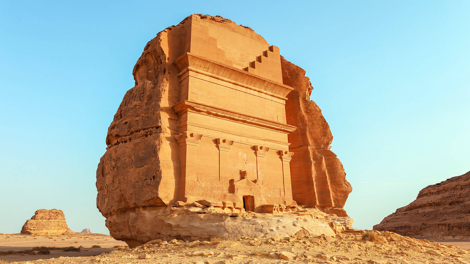 Saudi 桌布 壁紙 — 一座雕刻在巨大砂岩巨石上的古老石廟，獨自矗立在廣闊的沙漠中，天空湛藍清澈。