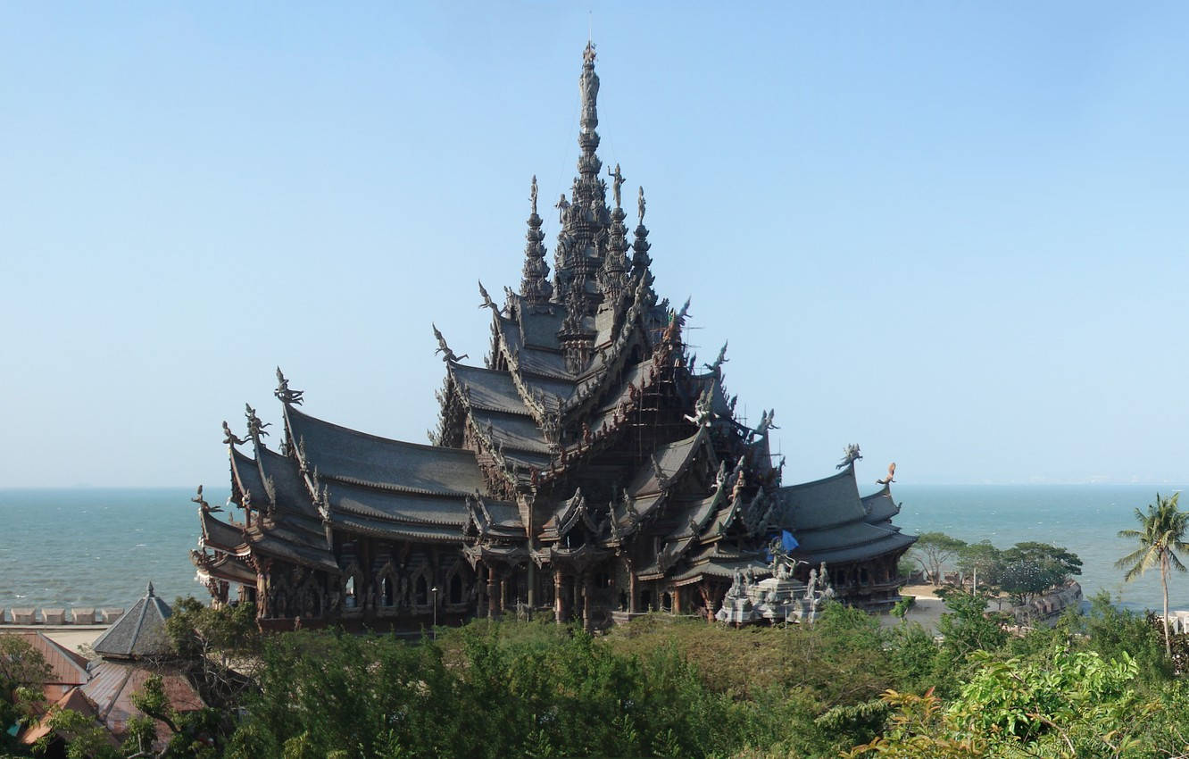 Architecture 桌布 壁紙 — 一座雕刻精緻的深色木造寺廟，有多個尖塔，在清澈的藍天下，雄偉地聳立於海洋旁茂密的綠植之上。