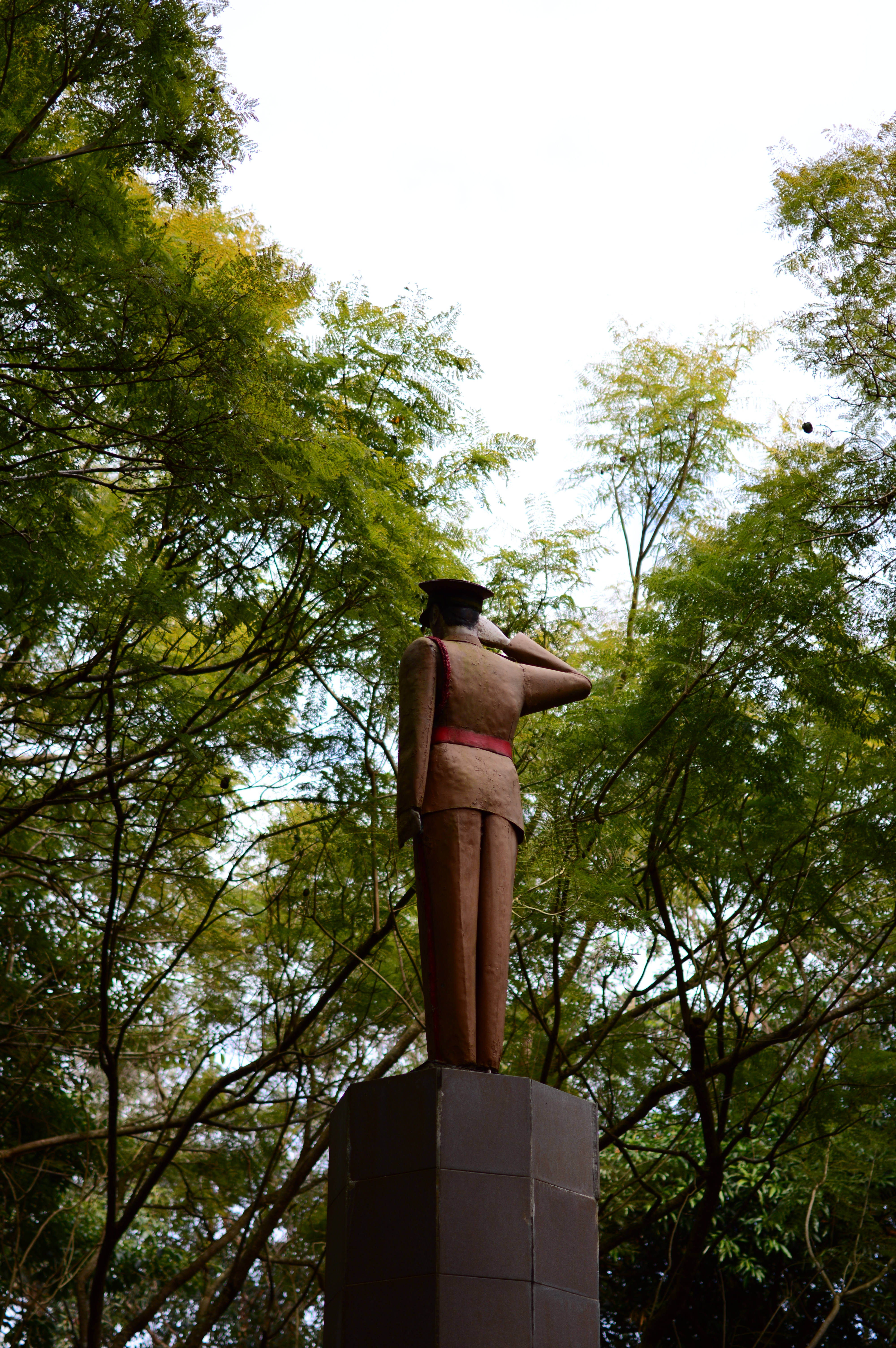 Mauritius 桌布 壁紙 — 一座青銅士兵雕像在基座上敬禮，周圍環繞著茂密的綠樹，天空明亮。