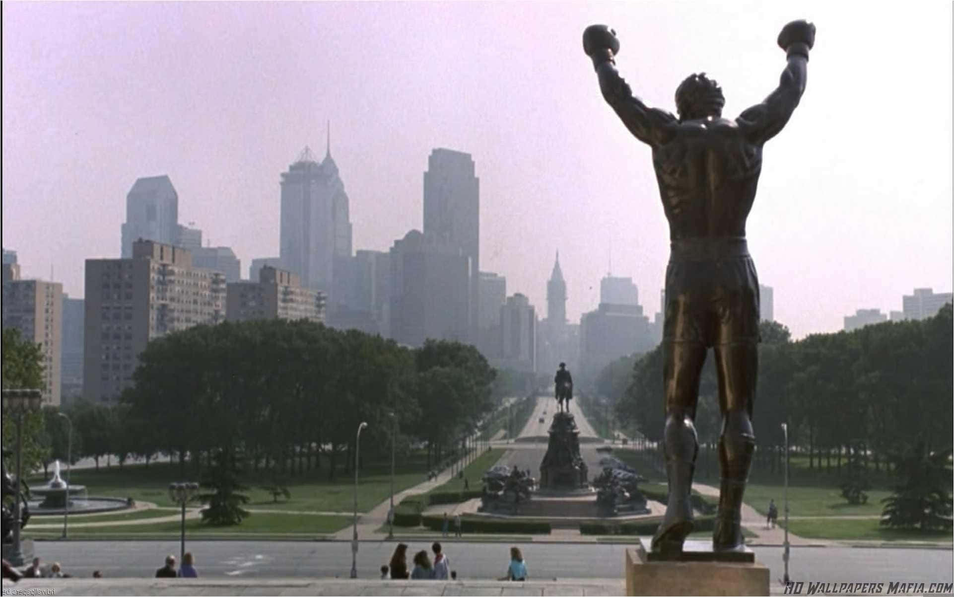 Rocky Balboa 桌布 壁紙 — 一座青銅雕像，描繪一位勝利的拳擊手高舉拳頭，矗立在公園中，面向朦朧的城市天際線。
