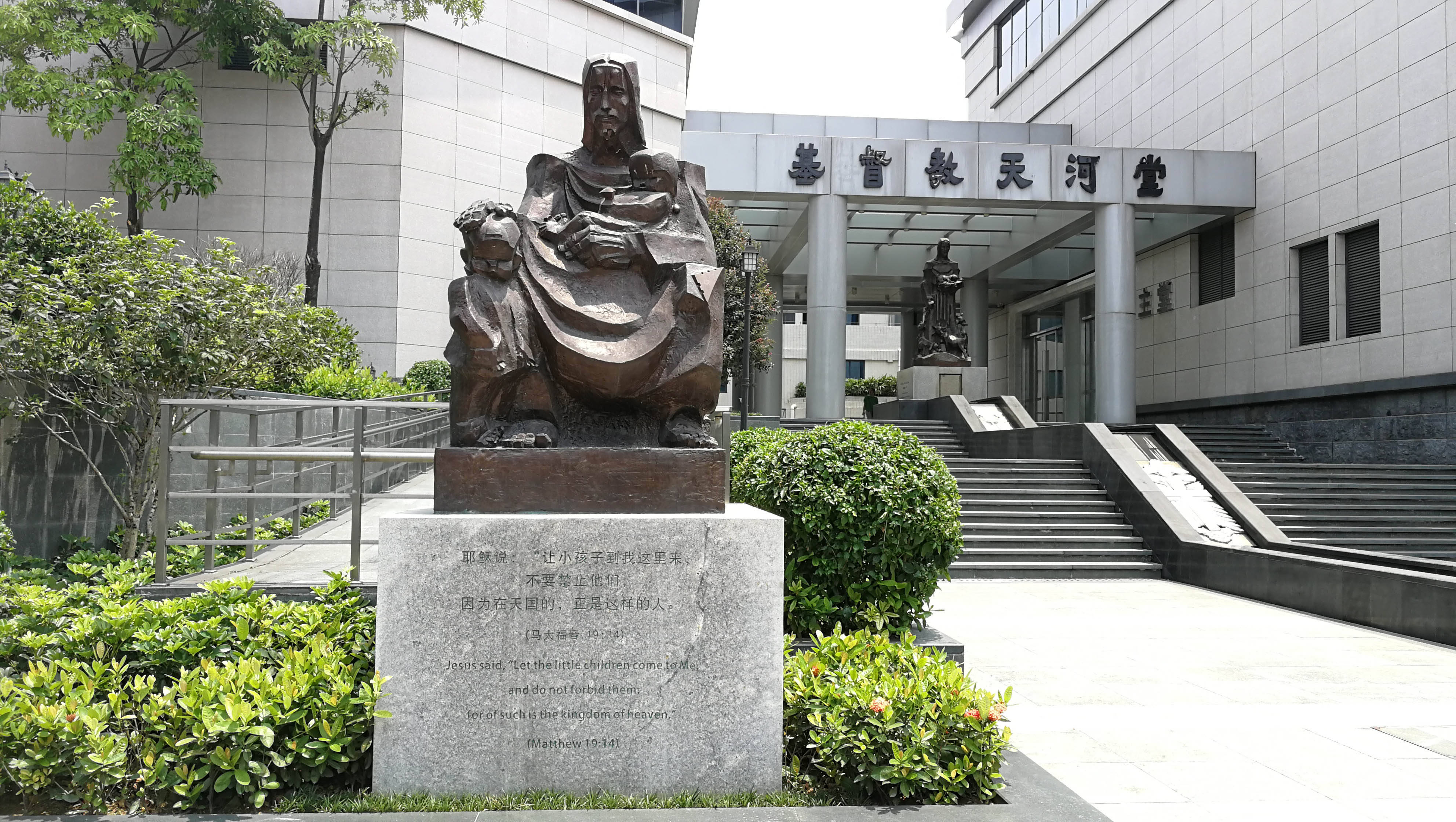 Guangzhou 桌布 壁紙 — 一座青銅雕像，描繪一位坐著的人物抱著一個孩子，矗立在一棟現代建築入口前，周圍環繞著綠色灌木和石階。
