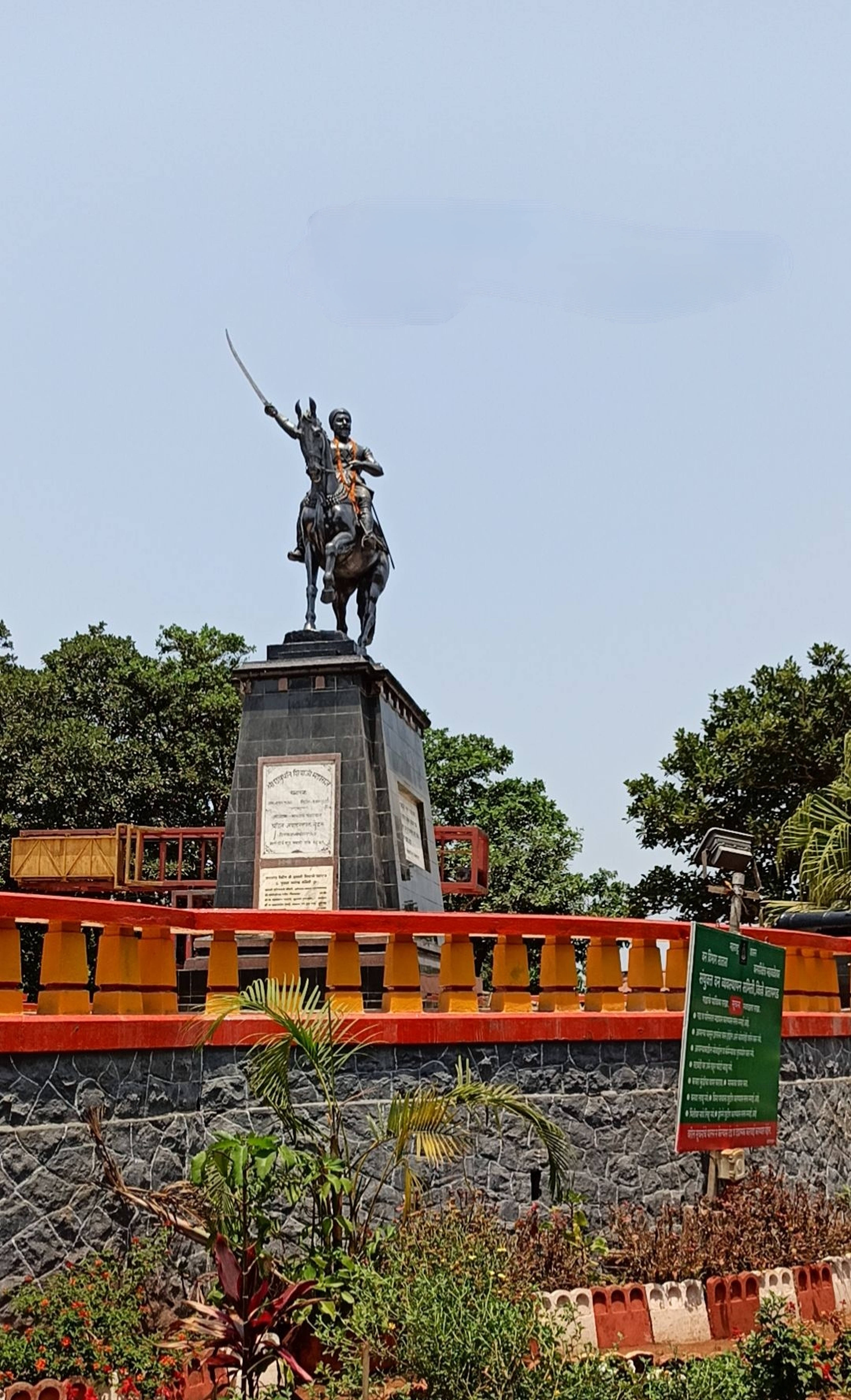 Indian 桌布 壁紙 — 一座青銅騎馬戰士雕像，手持劍，矗立在石質基座上，周圍環繞著茂密的綠樹和色彩繽紛的欄杆，天空晴朗。