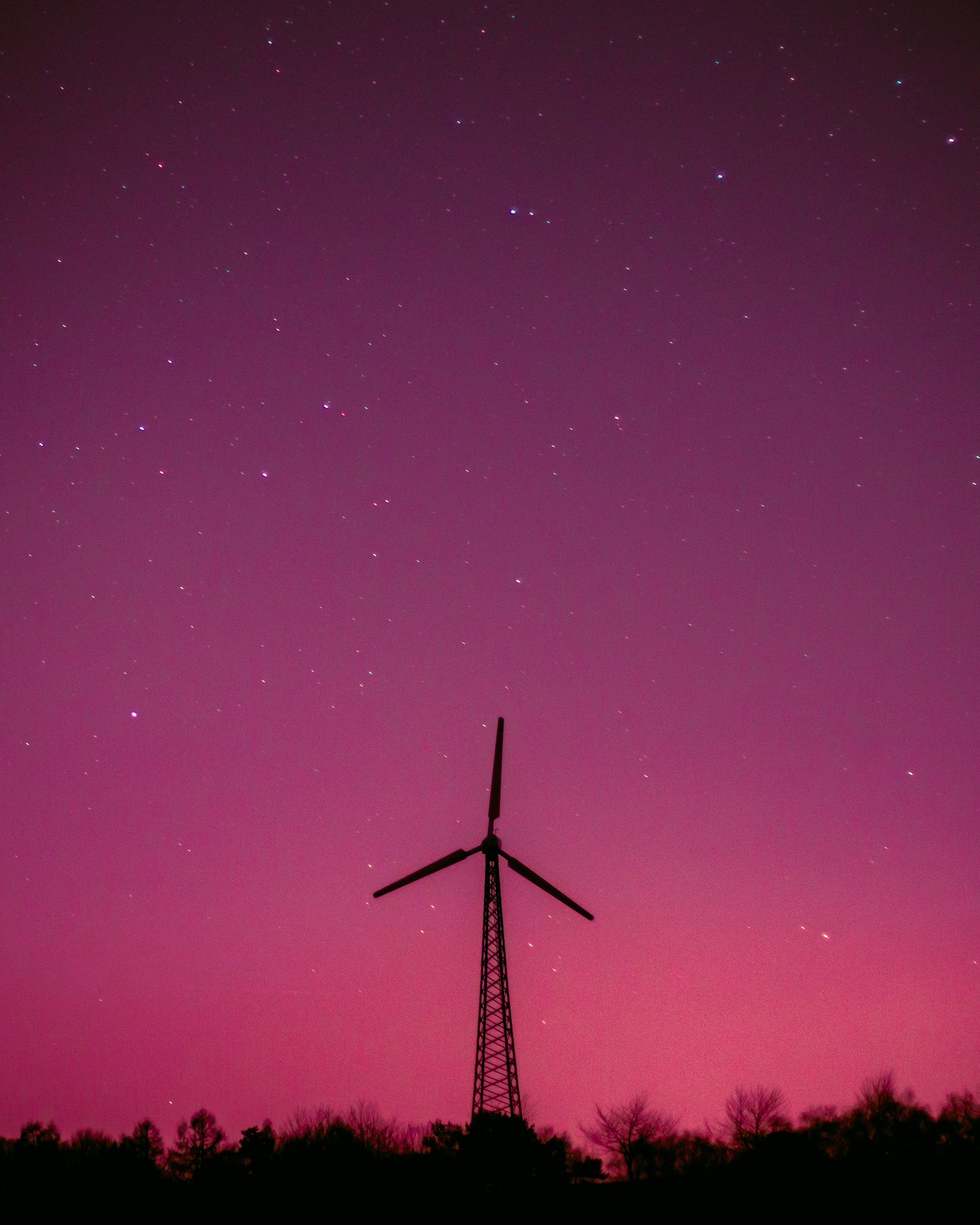 夜空 桌布 壁紙 — 一座風力渦輪機在樹線上方，以鮮豔的洋紅色星空夜空為背景，呈現出黑暗的輪廓。