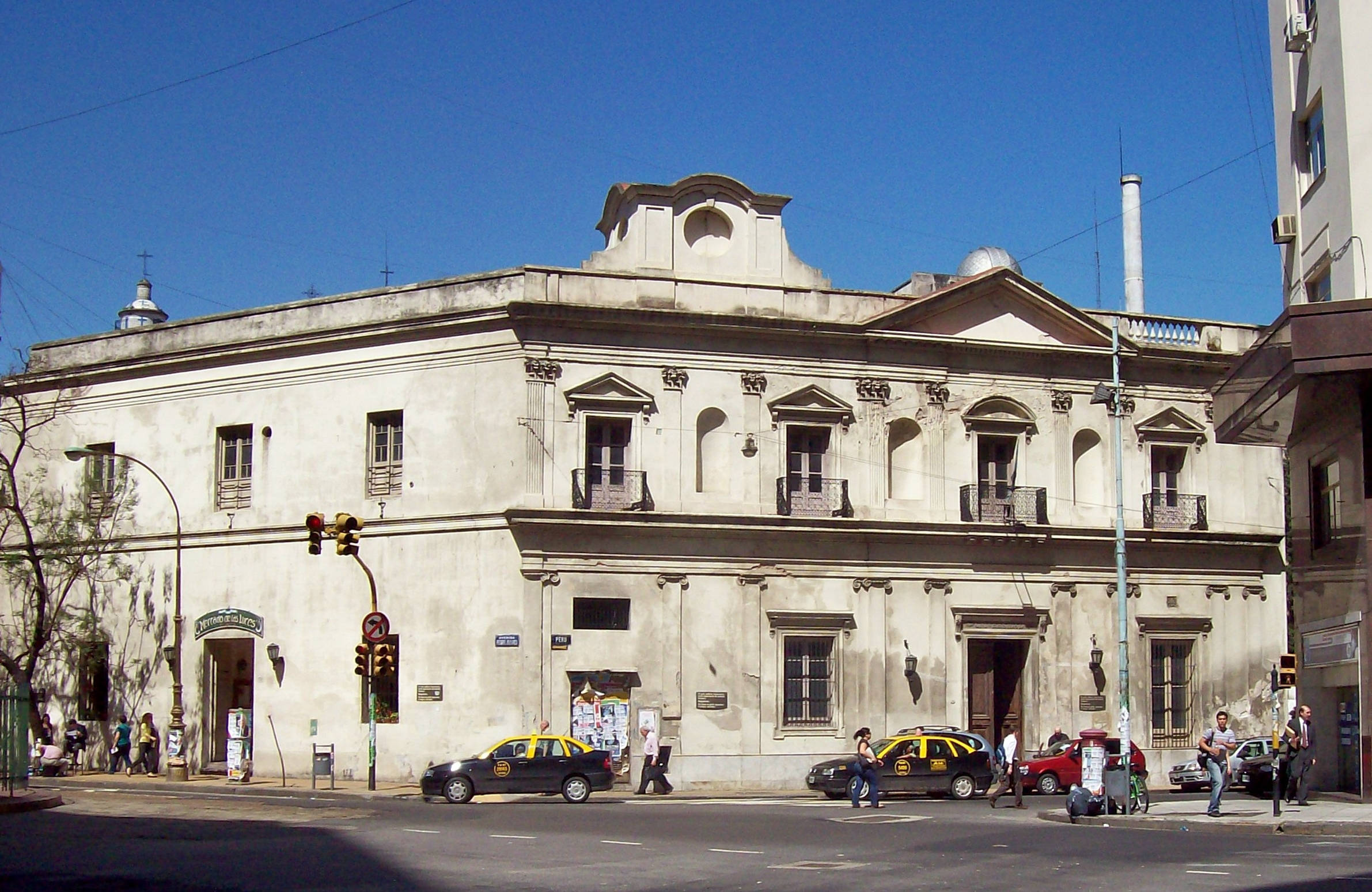 Aires 桌布 壁紙 — 一座風化的新古典主義建築，帶有拱形窗戶和陽台，矗立在晴朗的藍天下，計程車和行人沿著街道移動。
