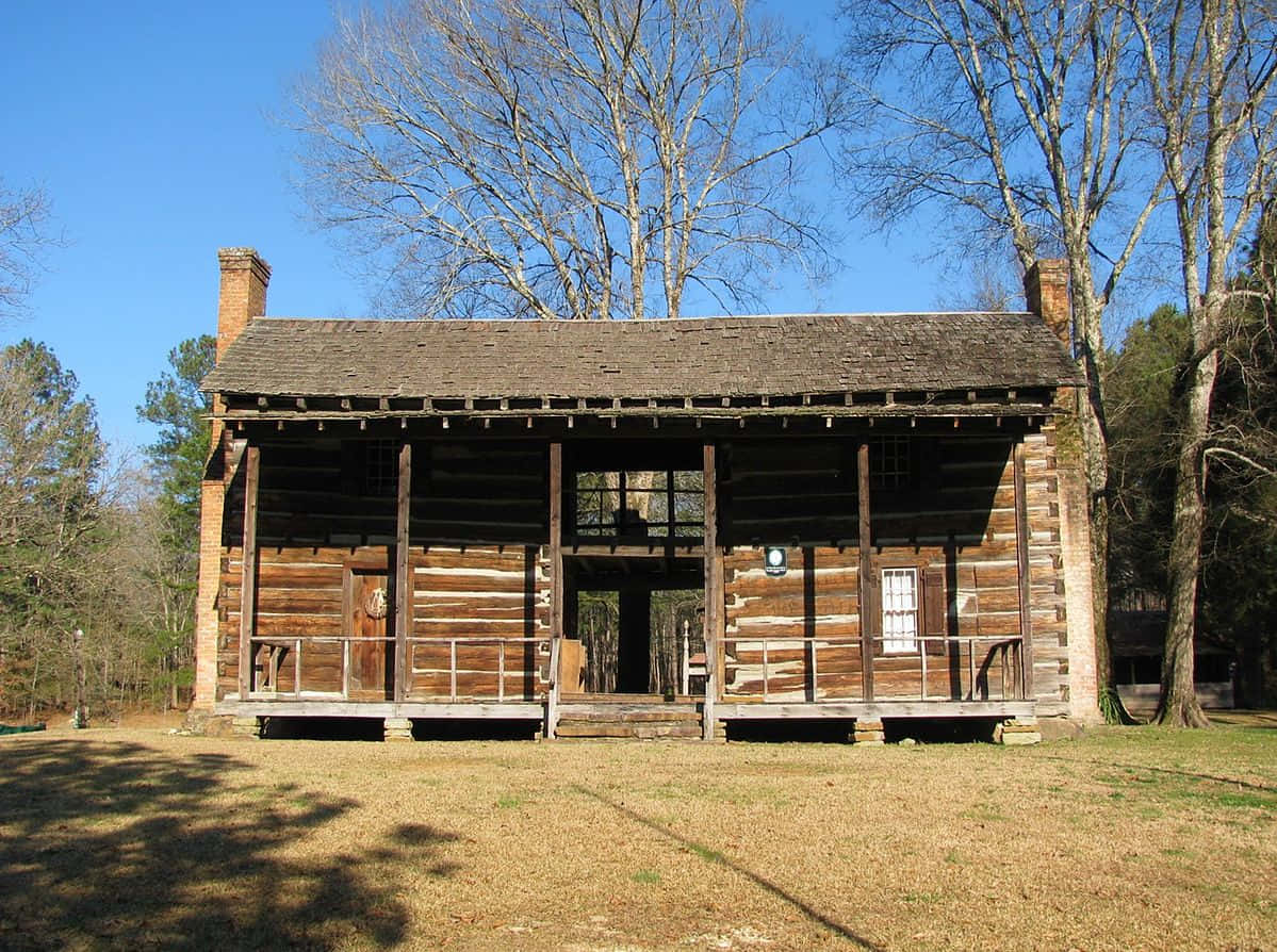 Log Cabin 桌布 壁紙 — 一座風化的木屋，帶有寬闊的門廊，矗立在晴朗的藍天下，周圍是光禿禿的樹木和乾草。