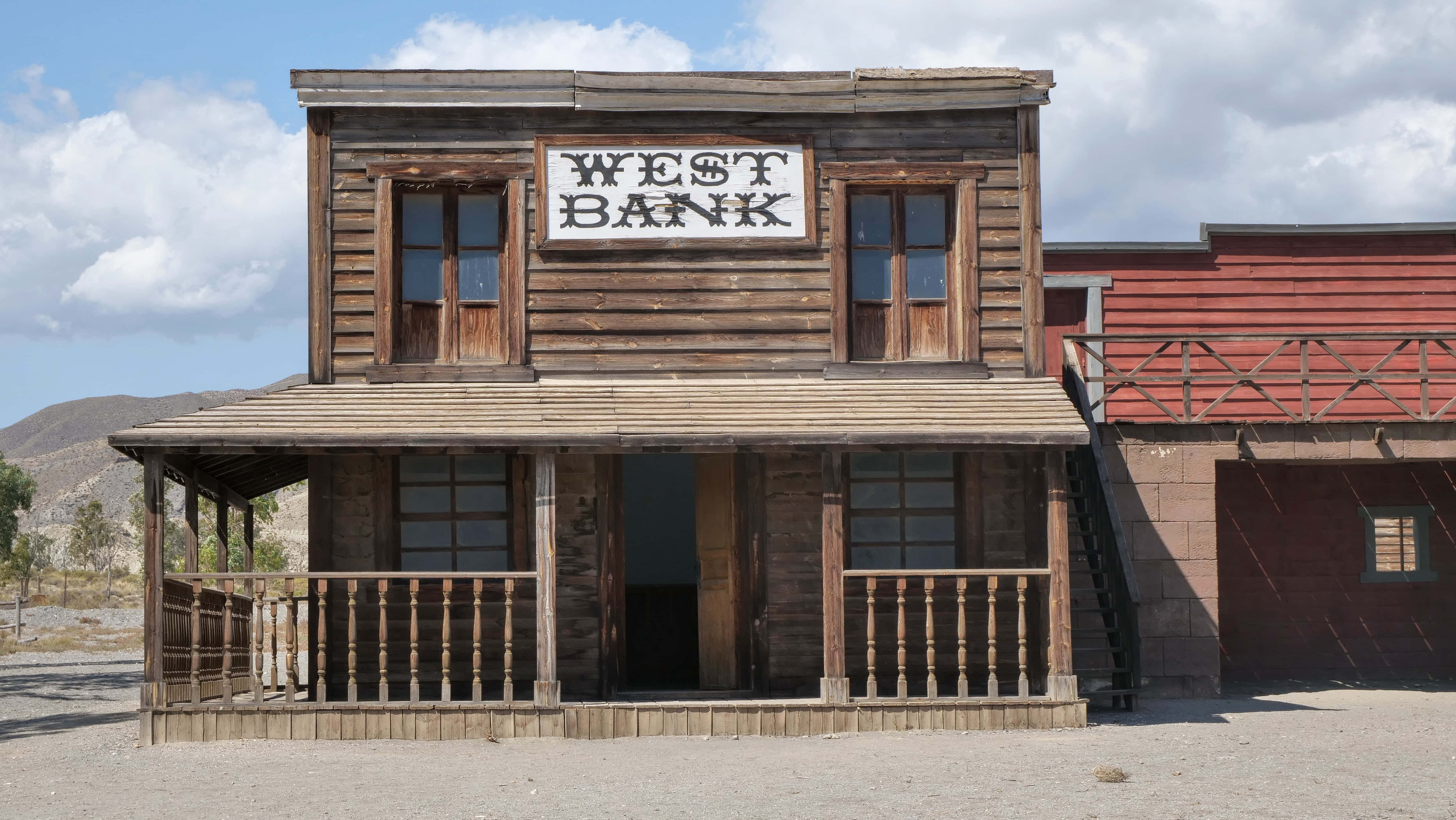 西部 桌布 壁紙 — 一座風化的木製兩層銀行建築，帶有門廊和'WEST BANK'標誌，矗立在塵土飛揚的沙漠小鎮，天空部分多雲。