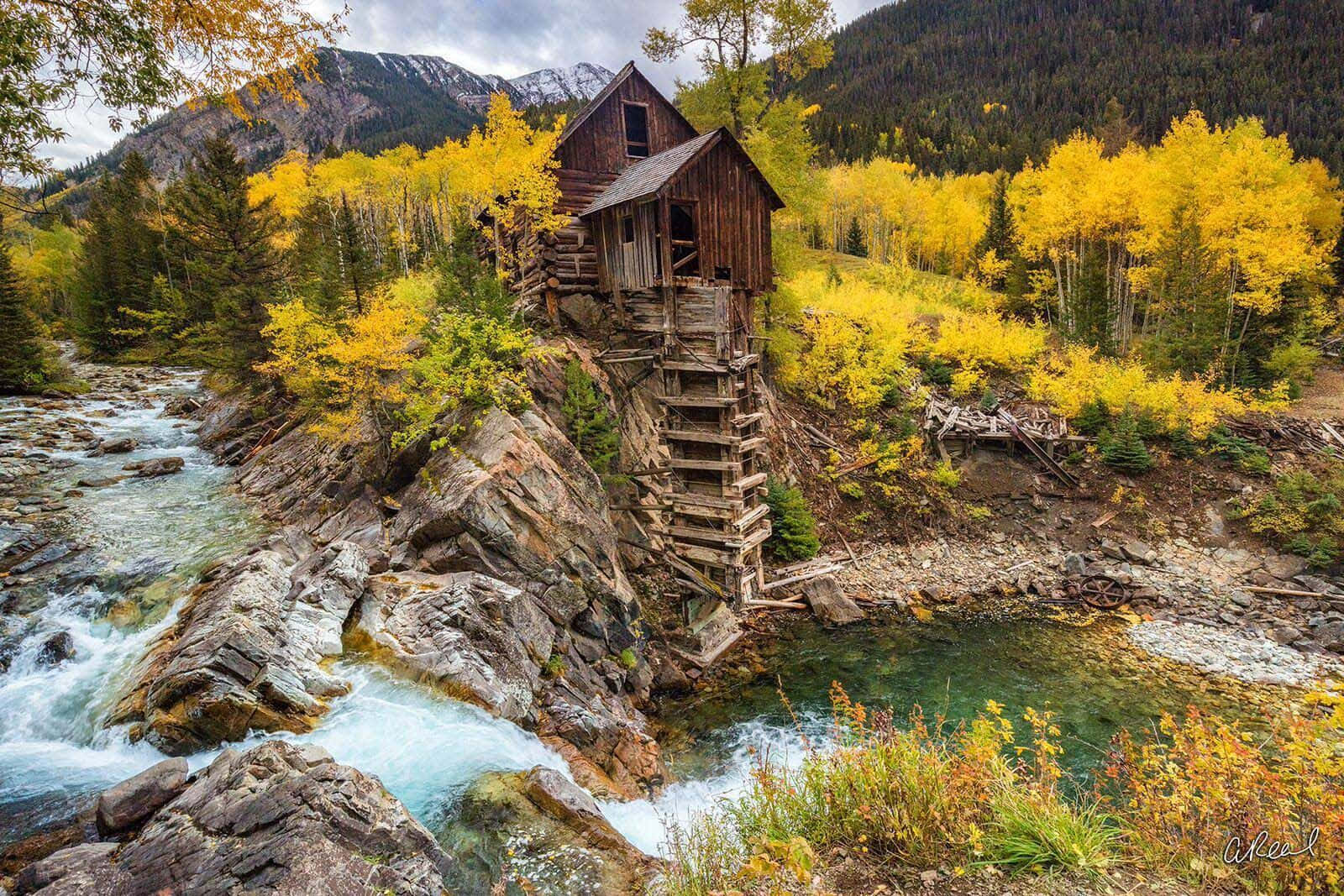 Nature Landscape 桌布 壁紙 — 一座風化的木製磨坊坐落在岩石河岸上，周圍環繞著鮮黃色的白楊樹和積雪覆蓋的山脈。