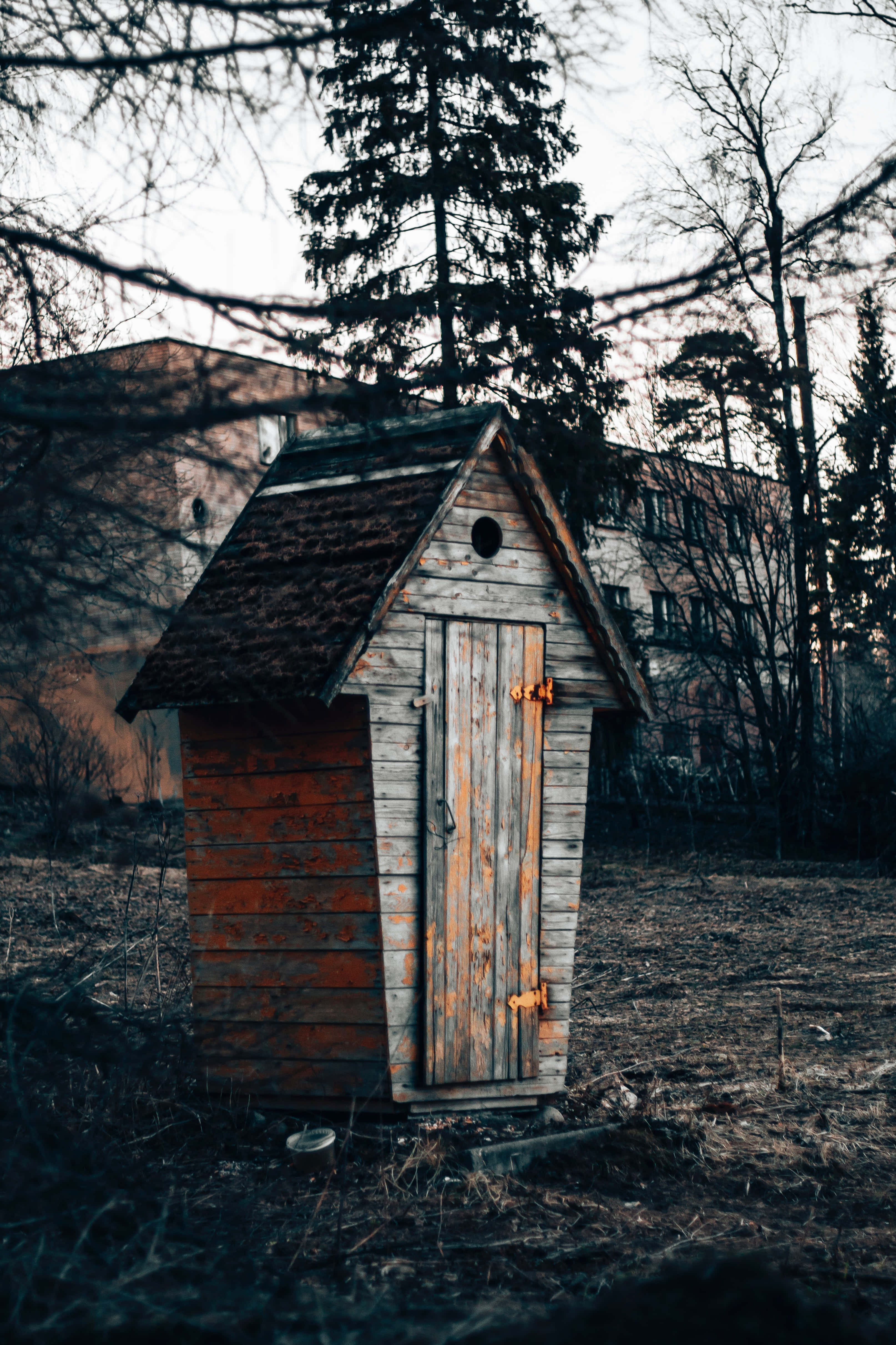 Outhouse 桌布 壁紙 — 一座風化的木造外屋孤零零地矗立在貧瘠的田野中，背景是光禿禿的樹木和褪色的建築物，天空陰沉。