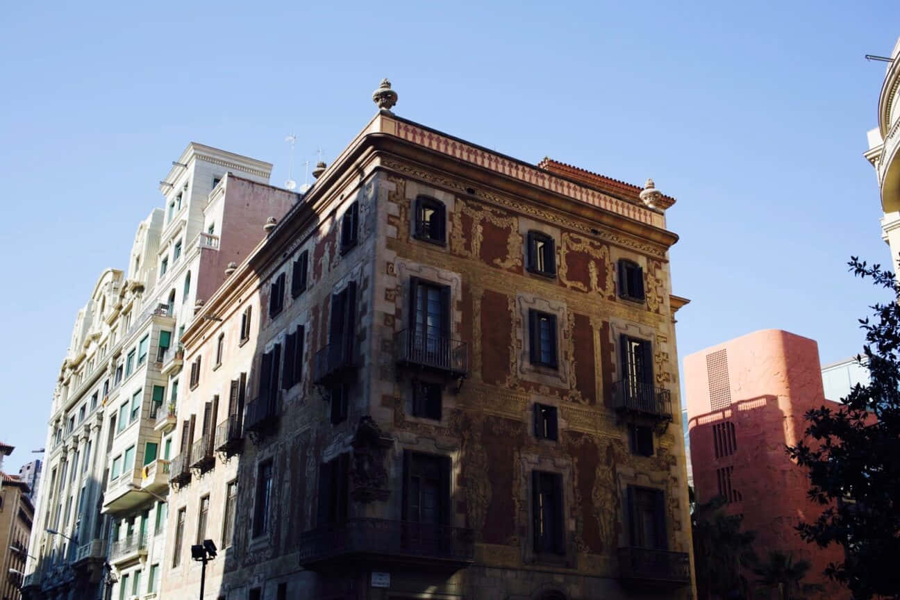 Barcelona 桌布 壁紙 — 一座風化的歷史建築，帶有華麗的陽台和褪色的壁畫，在晴朗的藍天下，矗立在陽光普照的城市環境中。