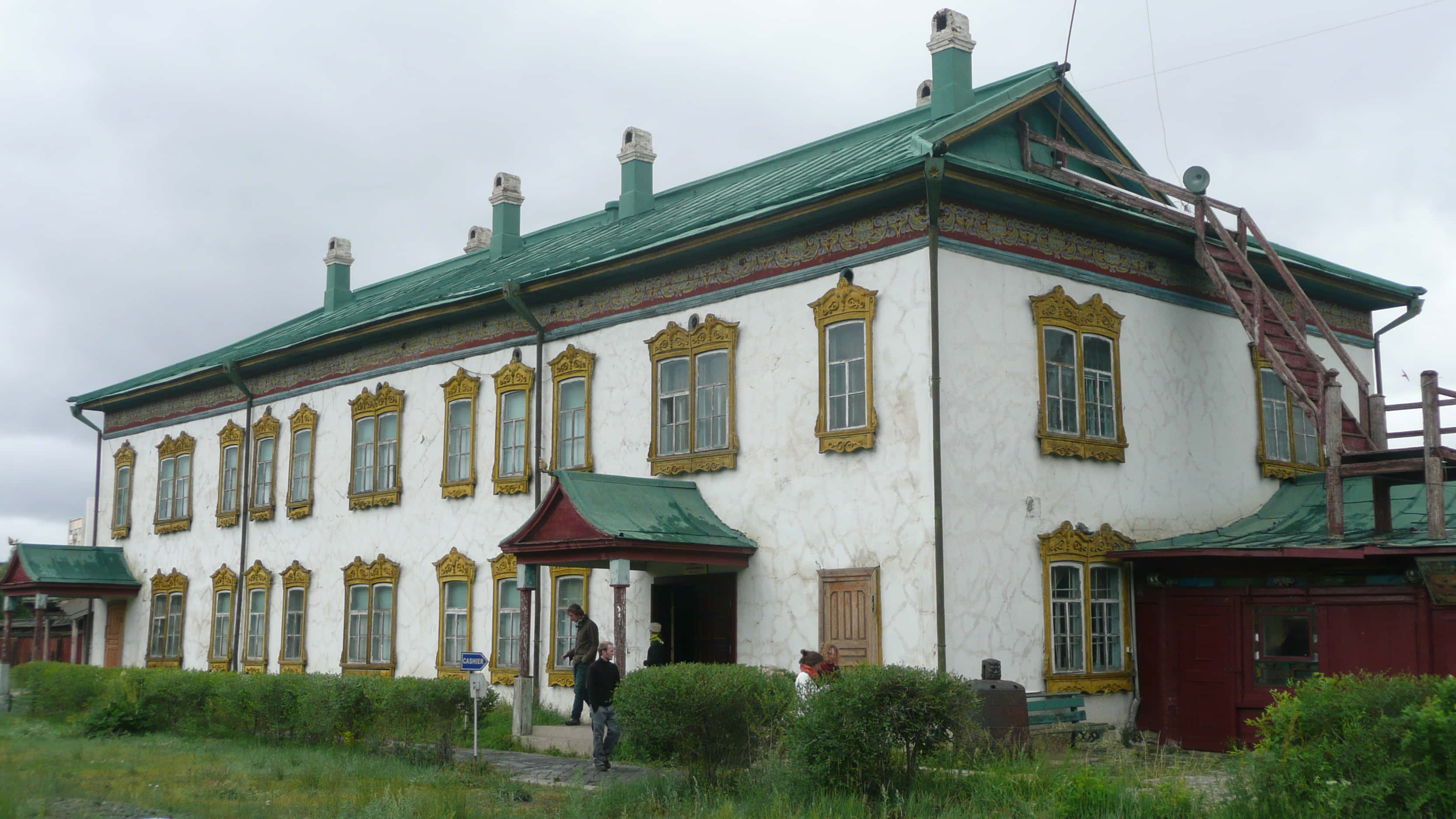 Mongolia 桌布 壁紙 — 一座風化的白色建築，帶有華麗的金色窗框和綠色屋頂，矗立在多雲的天空下，入口附近有人。