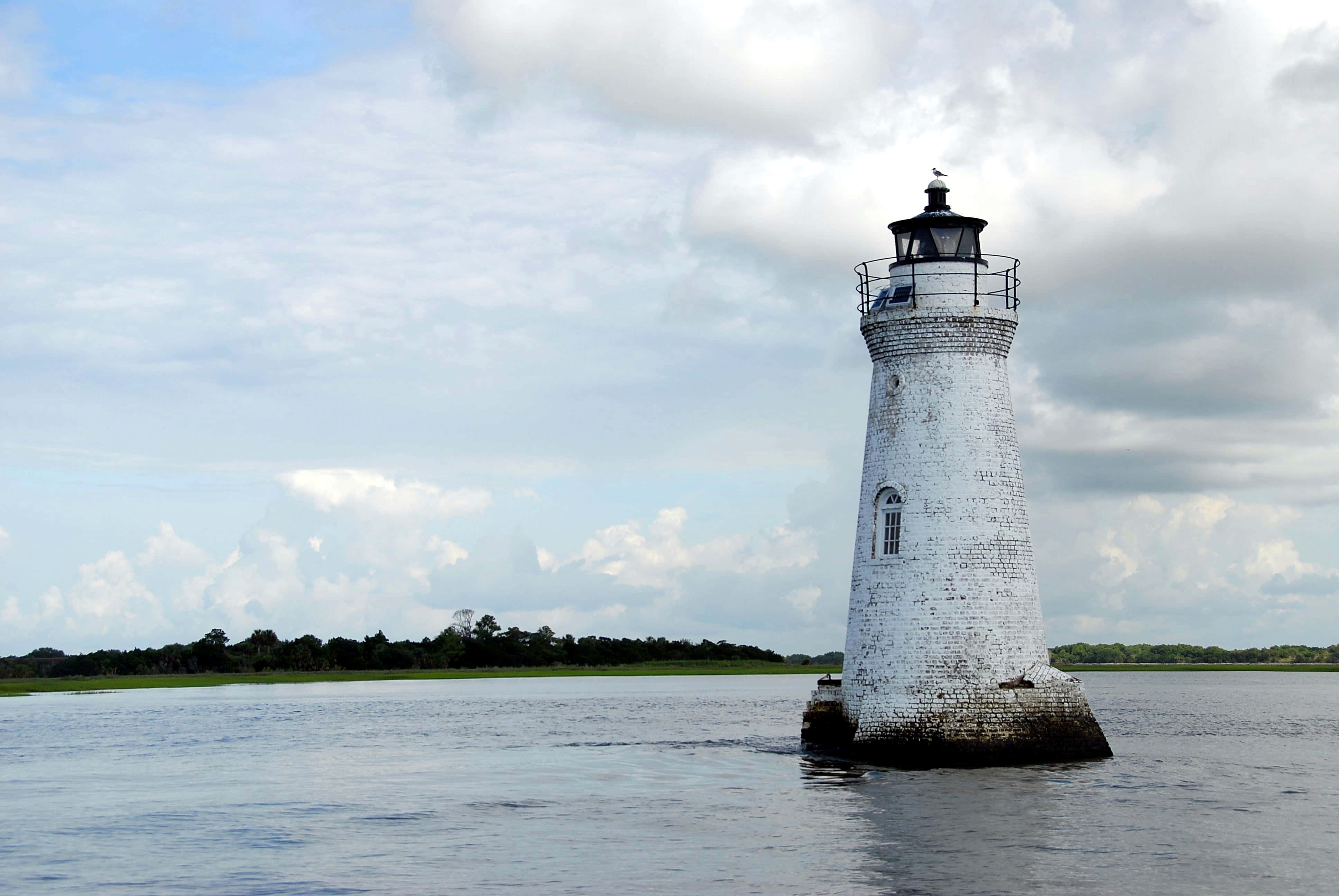 Lighthouse 桌布 壁紙 — 一座風化的白色磚砌燈塔孤獨地矗立在平靜的水面上，天空部分多雲。
