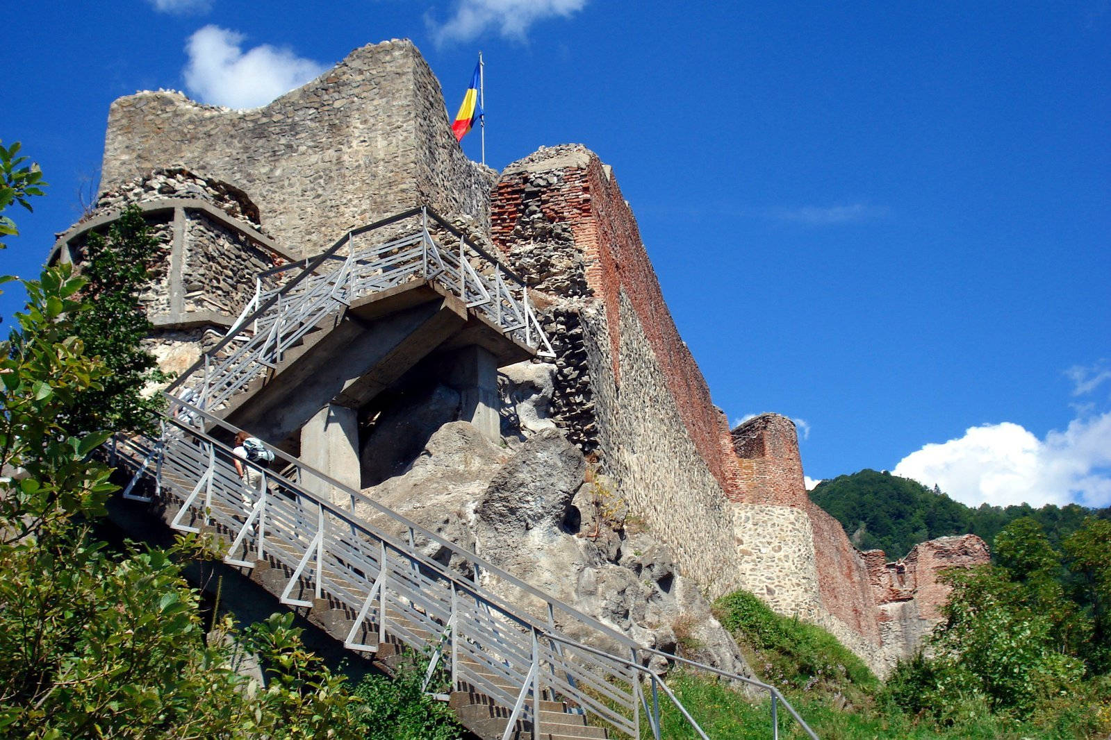 Romania 桌布 壁紙 — 一座風化的石城堡廢墟，現代金屬樓梯沿其岩石表面攀升，上方是晴朗藍天與零星白雲。