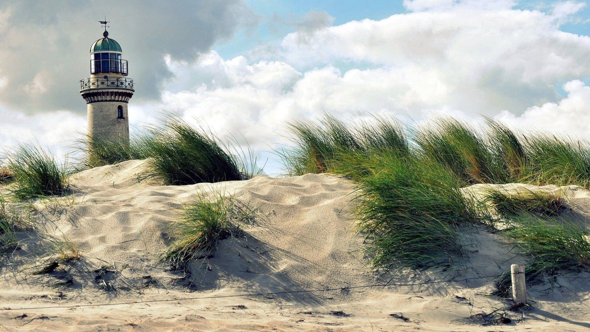 Beach Desktop 桌布 壁紙 — 一座風化的石燈塔矗立在沙丘上，高草在部分多雲的天空下搖曳。