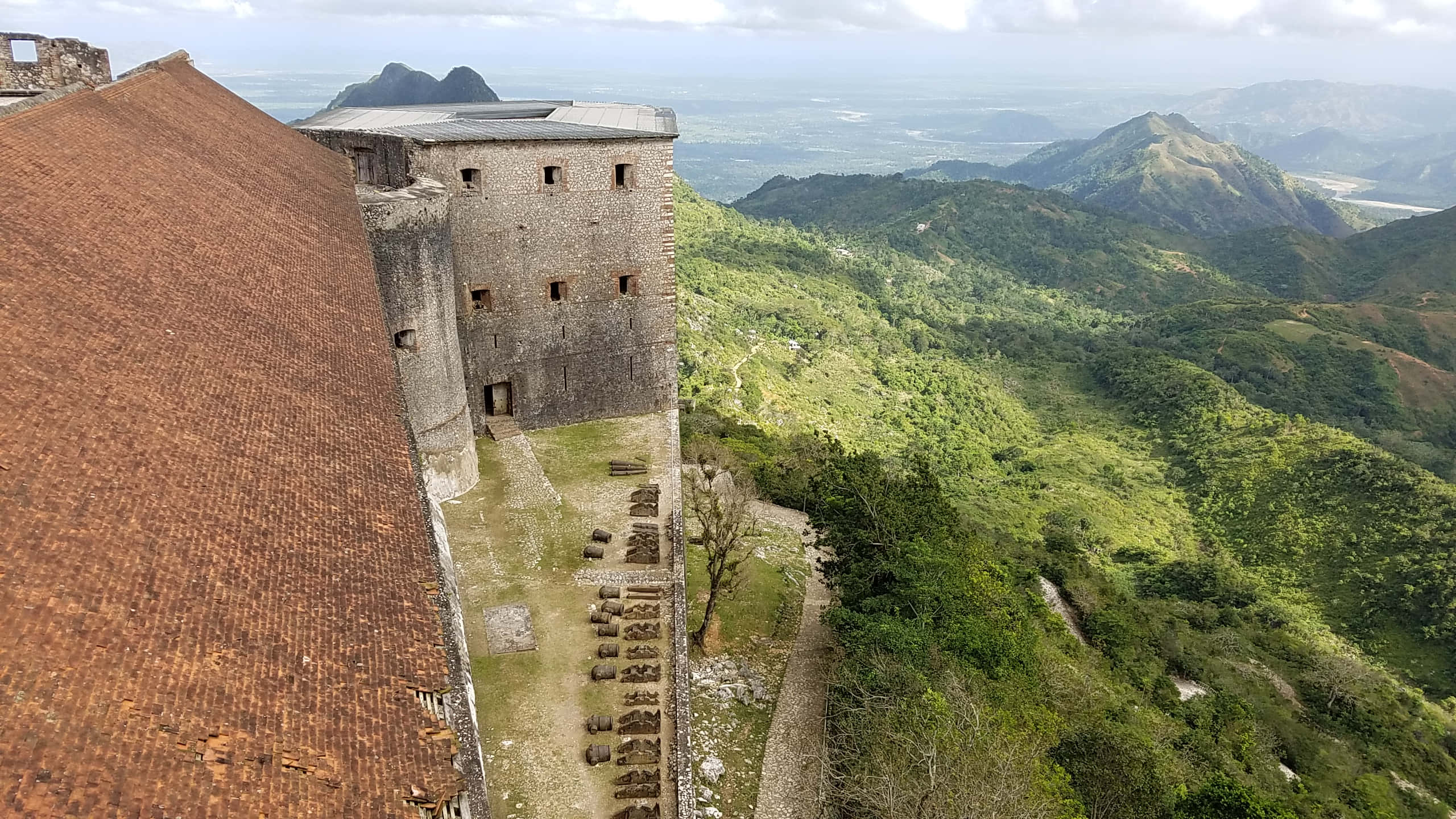 Fortress 桌布 壁紙 — 一座風化的石頭堡壘，帶有陶土屋頂，俯瞰著雲天下鬱鬱蔥蔥的山谷和遠山。