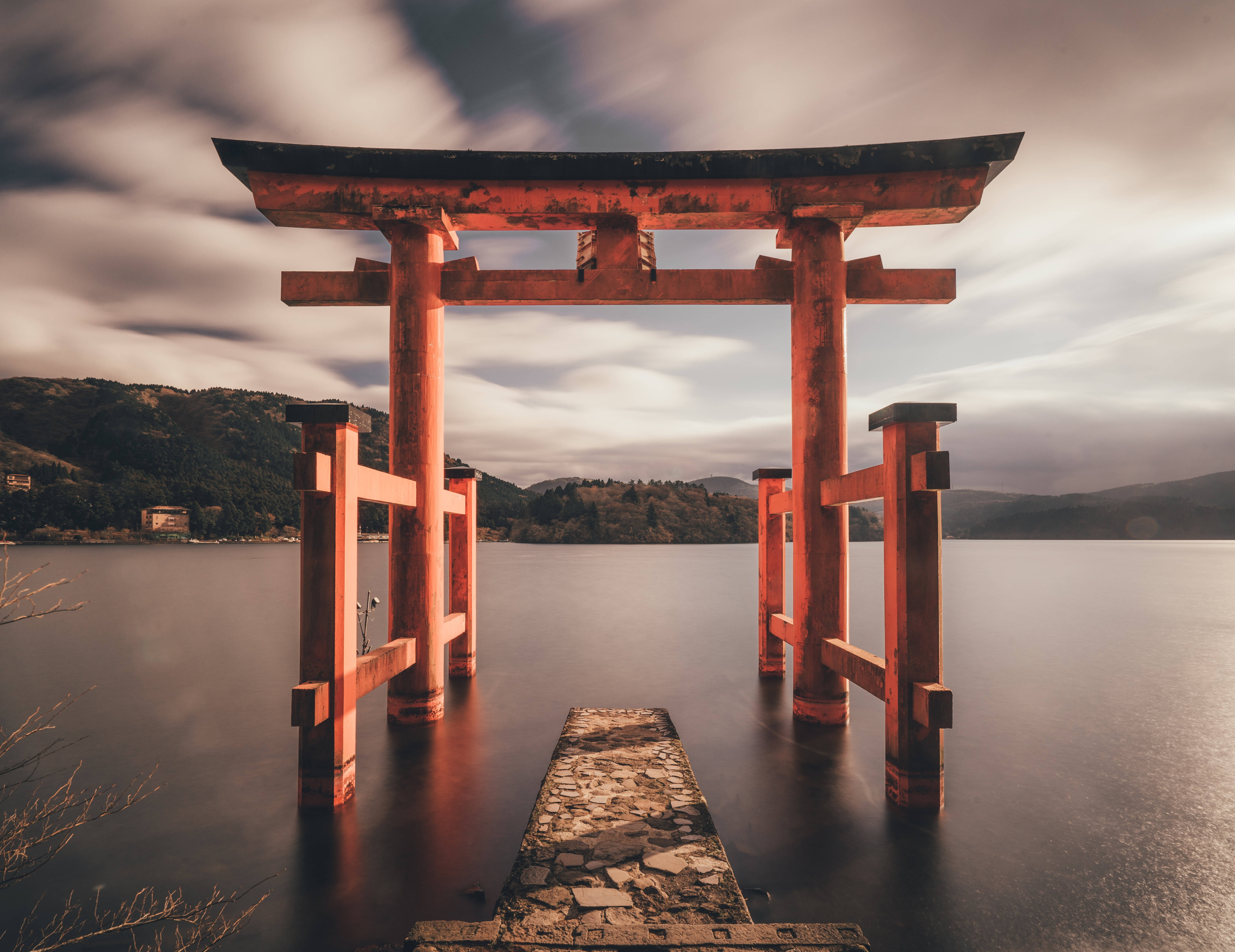 日本 桌布 壁紙 — 一座風化的紅色鳥居矗立在平靜的水面上，一條石徑在陰天下通往它。