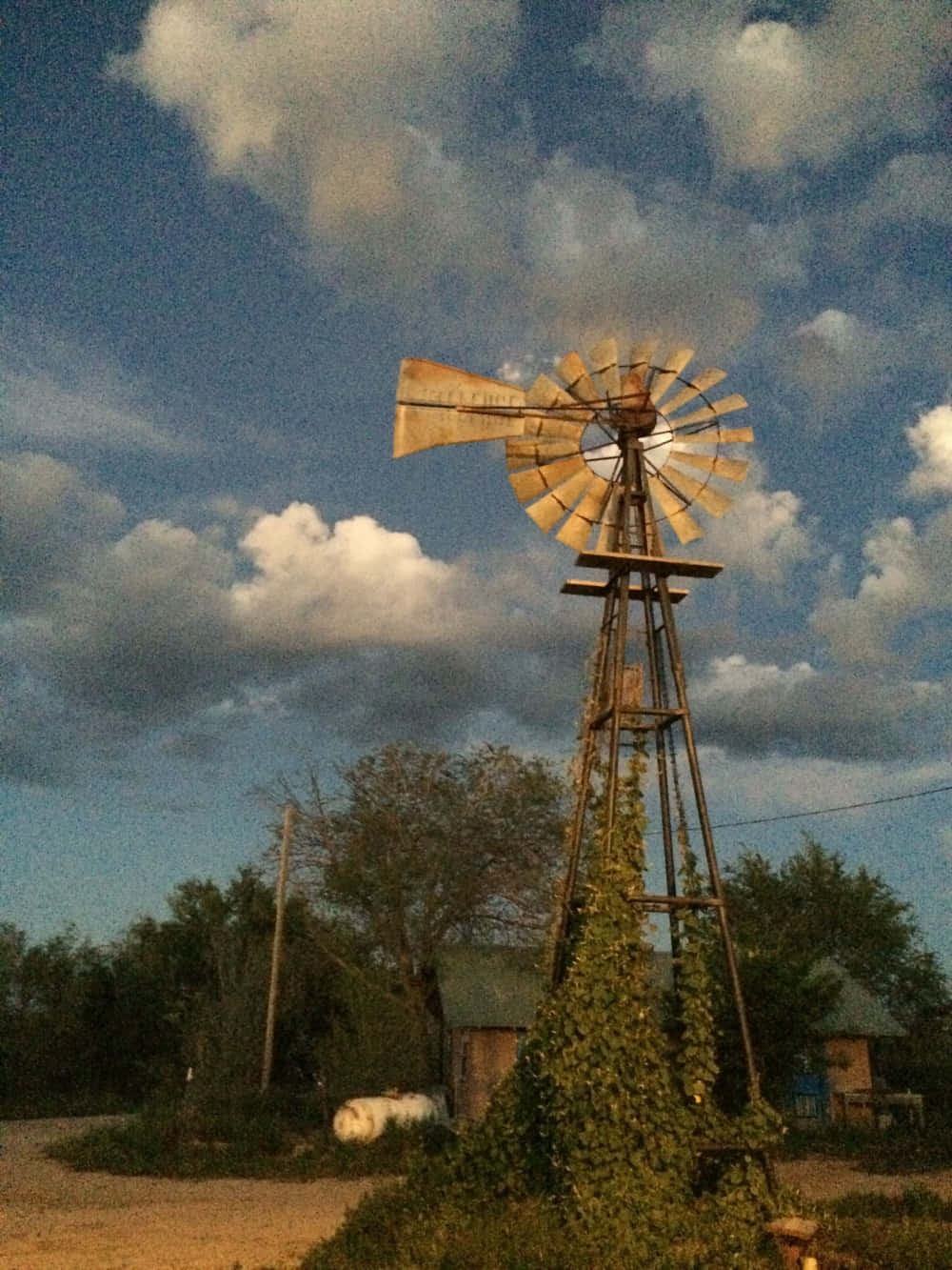 windmill 桌布 壁紙 — 一座風化的金屬風車在戲劇性的暮色天空下高聳矗立，天空中有散落的雲朵和周圍的樹木。