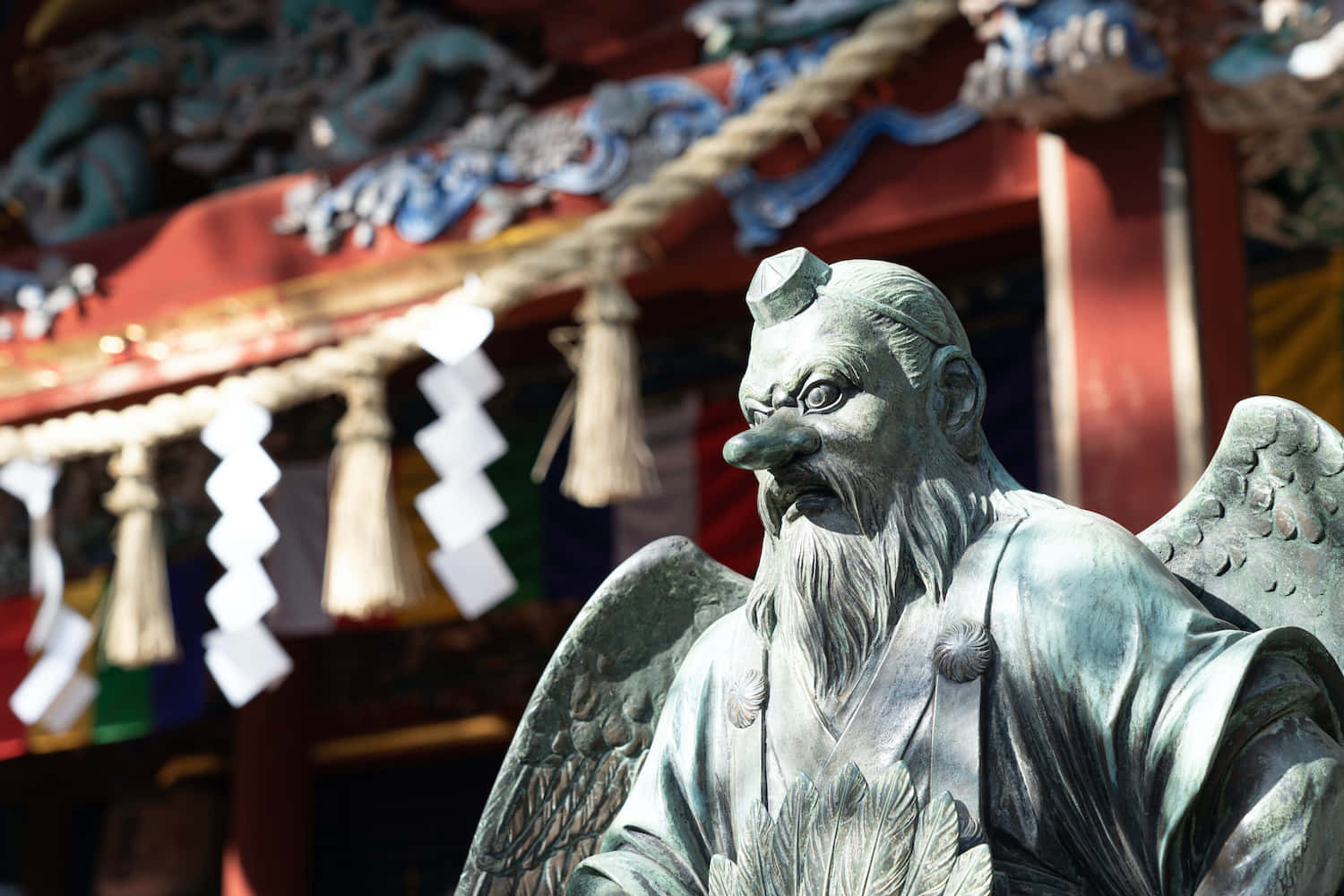 Tengu 桌布 壁紙 — 一座風化的青銅有翼鬍鬚守護者雕像，矗立在一個裝飾著色彩繽紛的鳥居和白色紙垂的充滿活力的日本神社前。