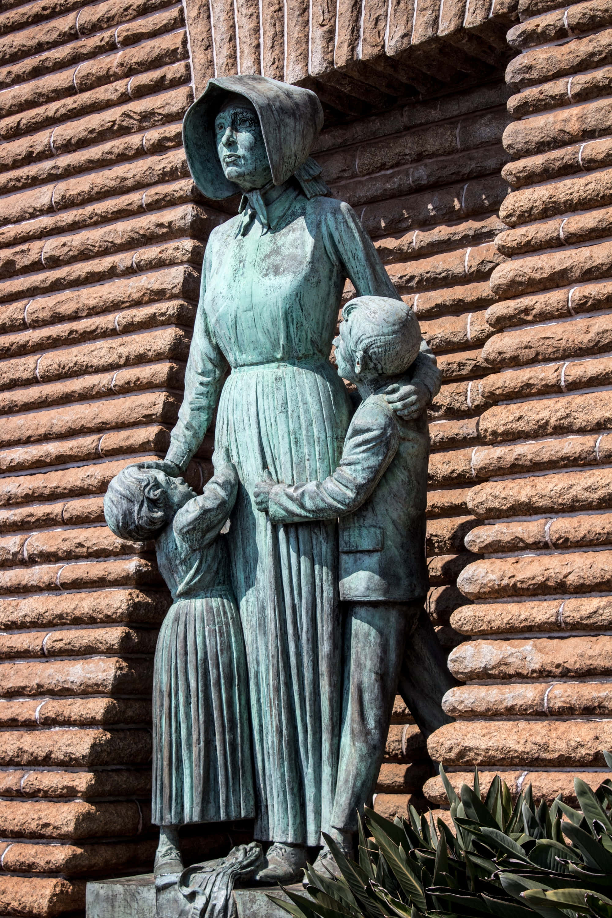 statue 桌布 壁紙 — 一座風化的青銅雕像，描繪一位女士和兩個孩子，倚著質感磚牆而立，底部有綠植。