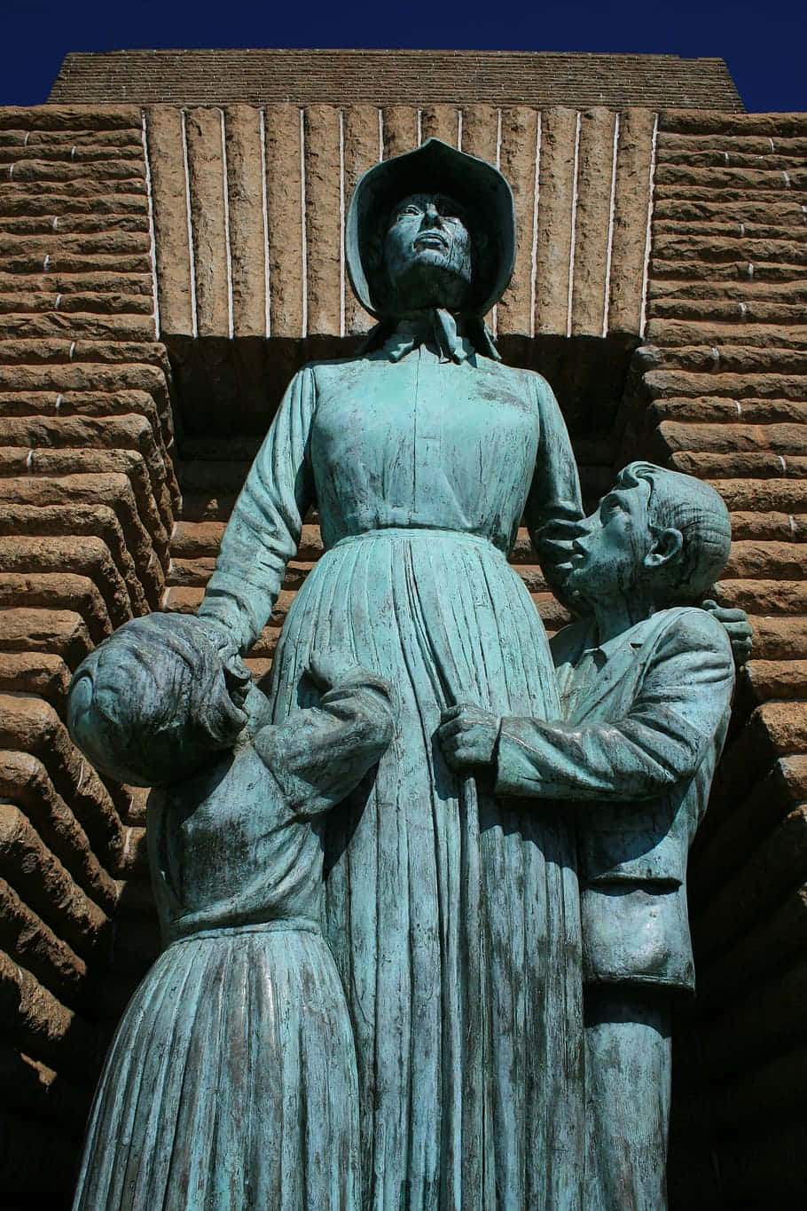 statue 桌布 壁紙 — 一座風化的青銅雕像，描繪一位女性和兩個孩子，矗立在紋理磚牆前，背景是晴朗的藍天。