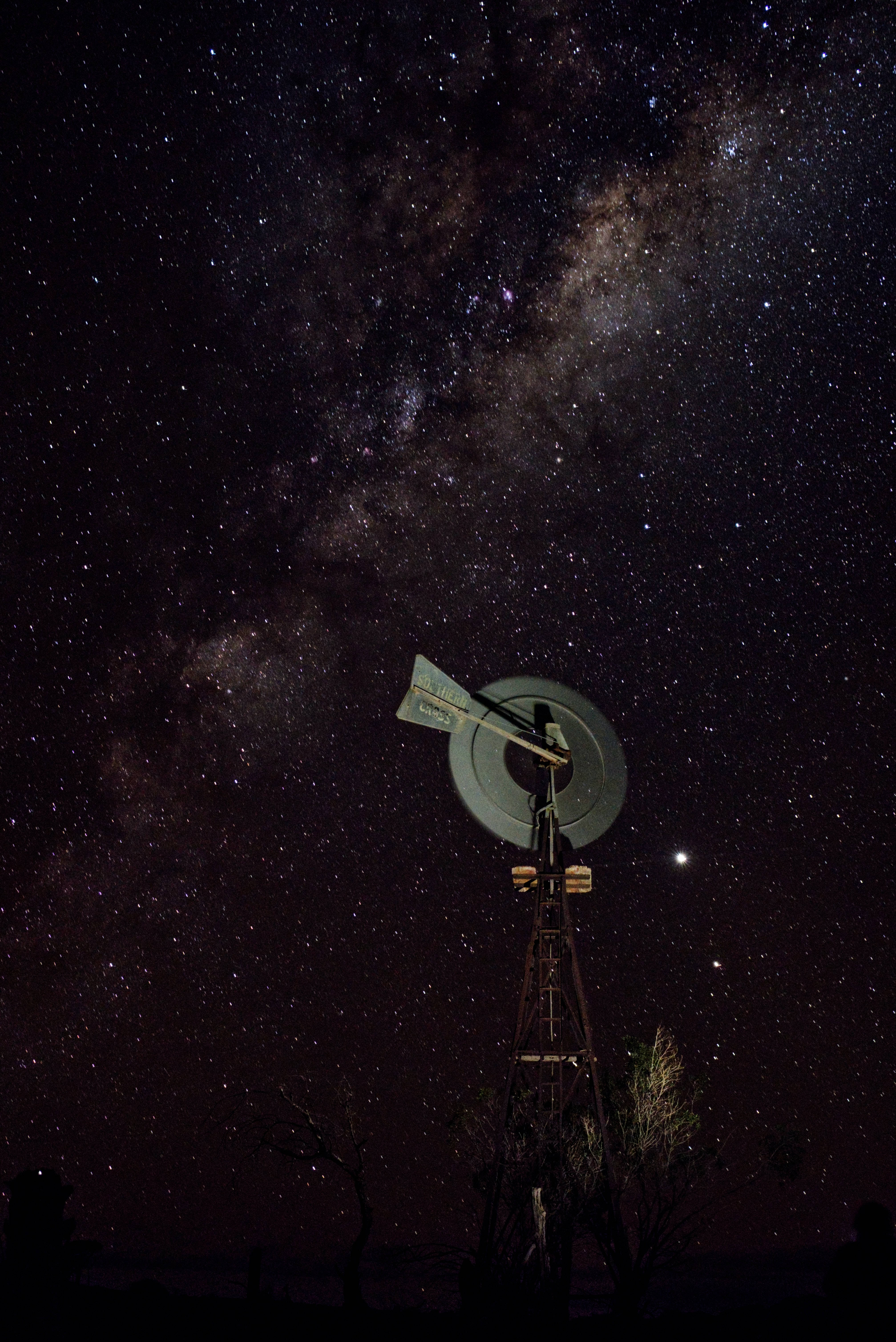 Starry 桌布 壁紙 — 一座飽經風霜的風車在壯麗的夜空下呈現剪影，夜空充滿了銀河系的密集星場和宇宙塵埃。