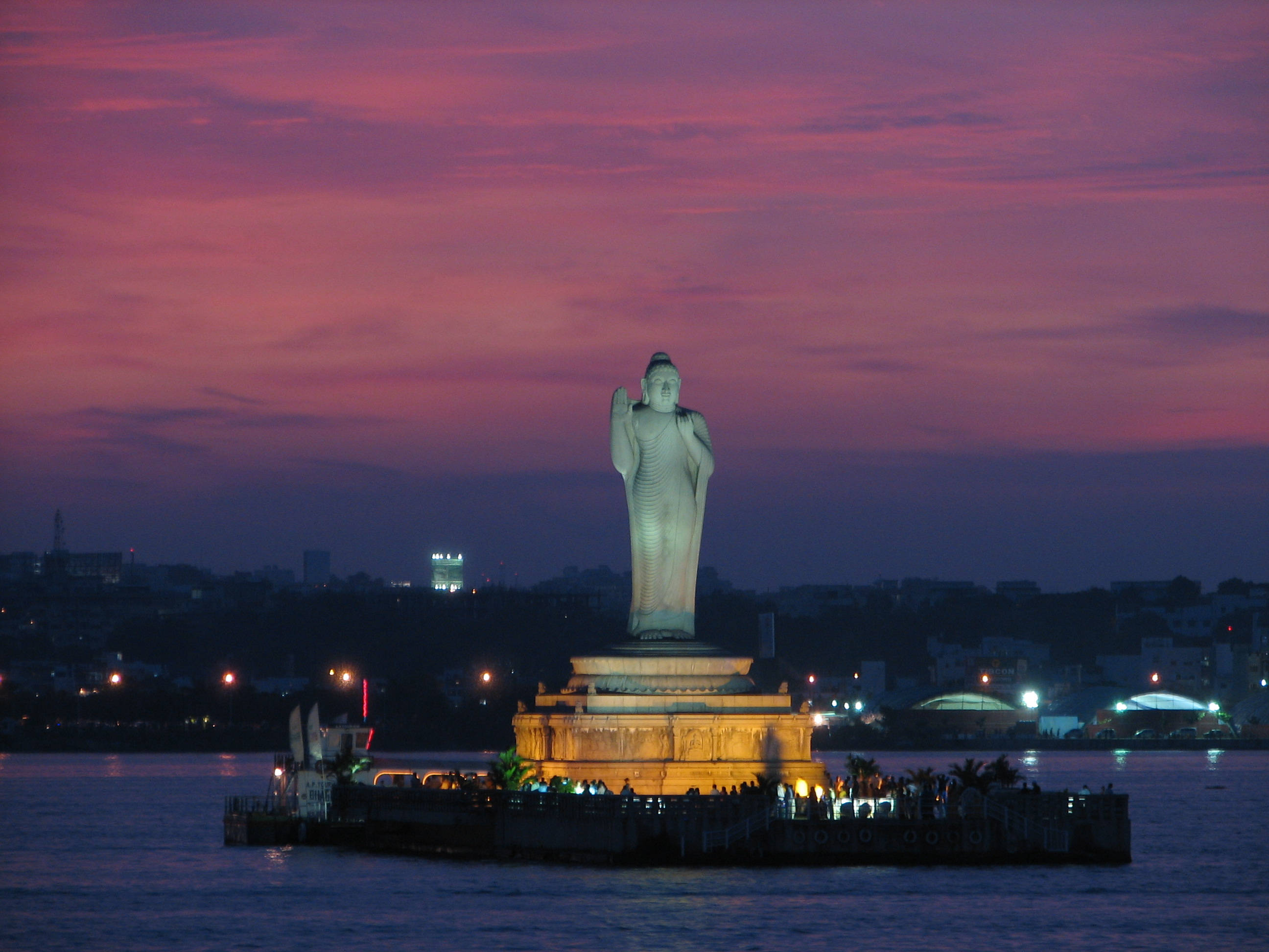 Hyderabad 桌布 壁紙 — 一座高大的佛像在充滿活力的粉紅色和紫色暮色天空下，在水濱平台上被照亮。
