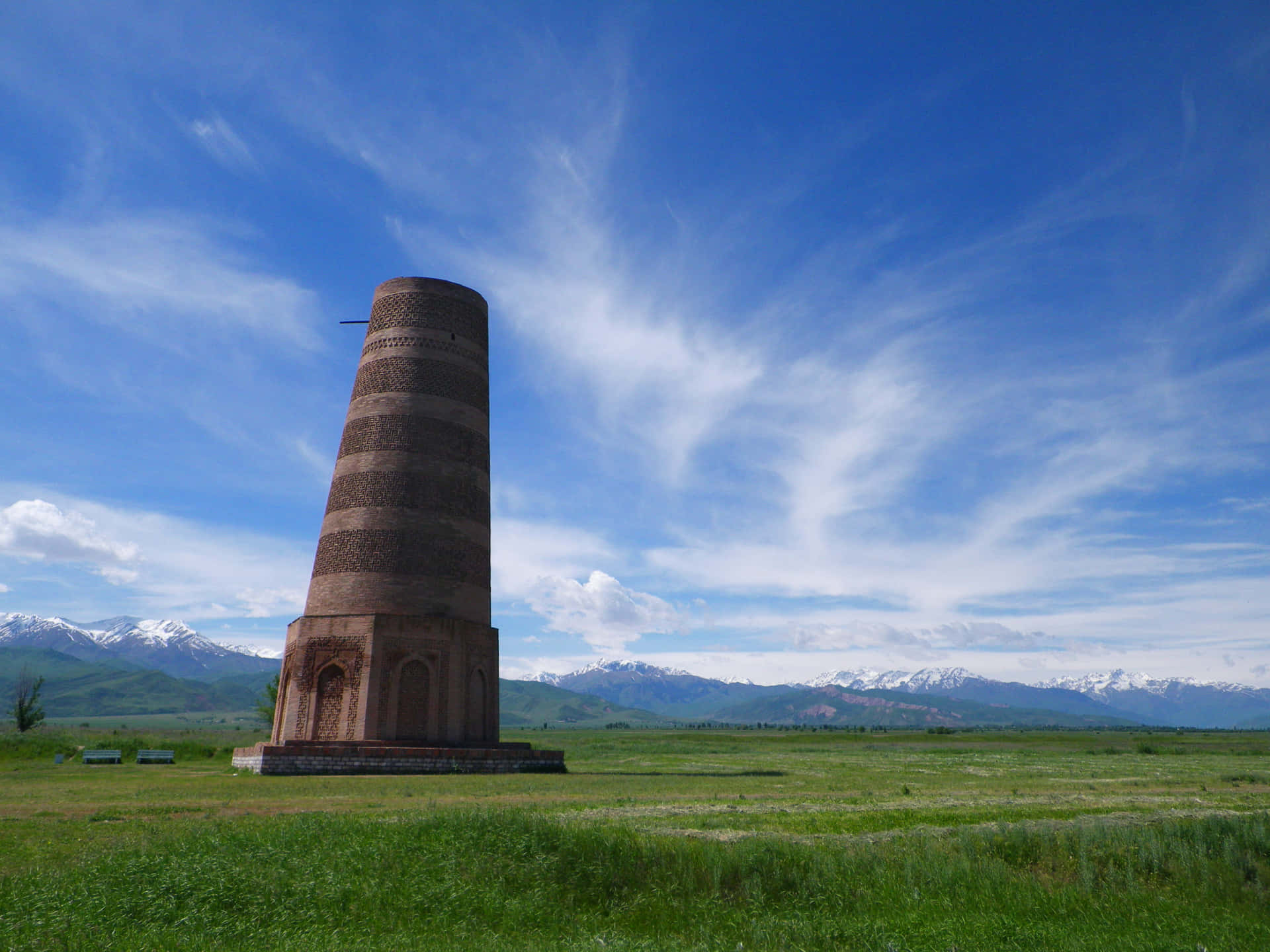 Kyrgyzstan 桌布 壁紙 — 一座高大的古老磚塔獨自矗立在廣闊的綠色田野中，天空湛藍，白雲縷縷，遠處有積雪覆蓋的山脈。