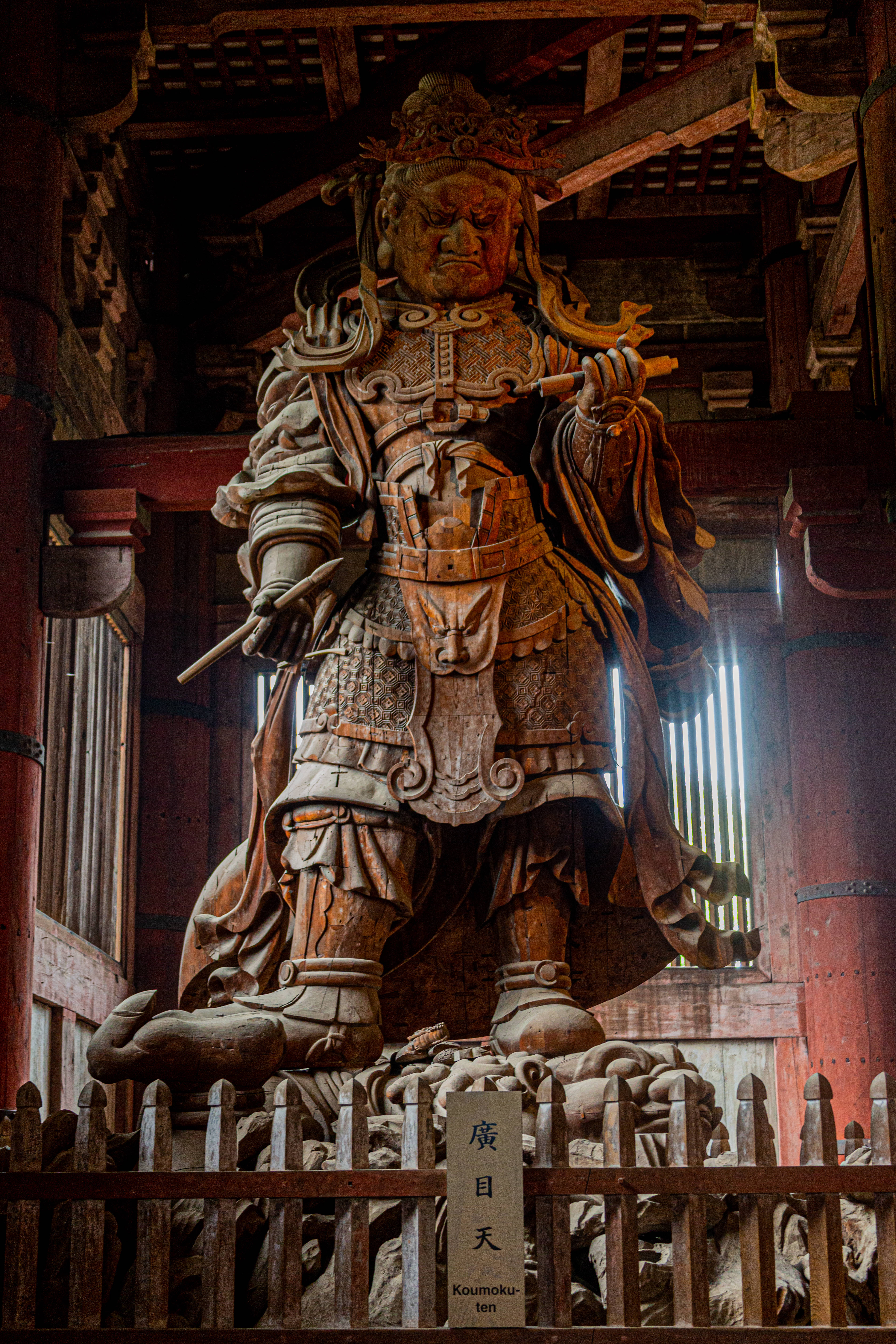 Warrior 桌布 壁紙 — 一座高大的木製廣目天像，身穿傳統日本盔甲的兇猛守護神，莊嚴地矗立在寺廟內，溫暖的木樑和光線照射其中。