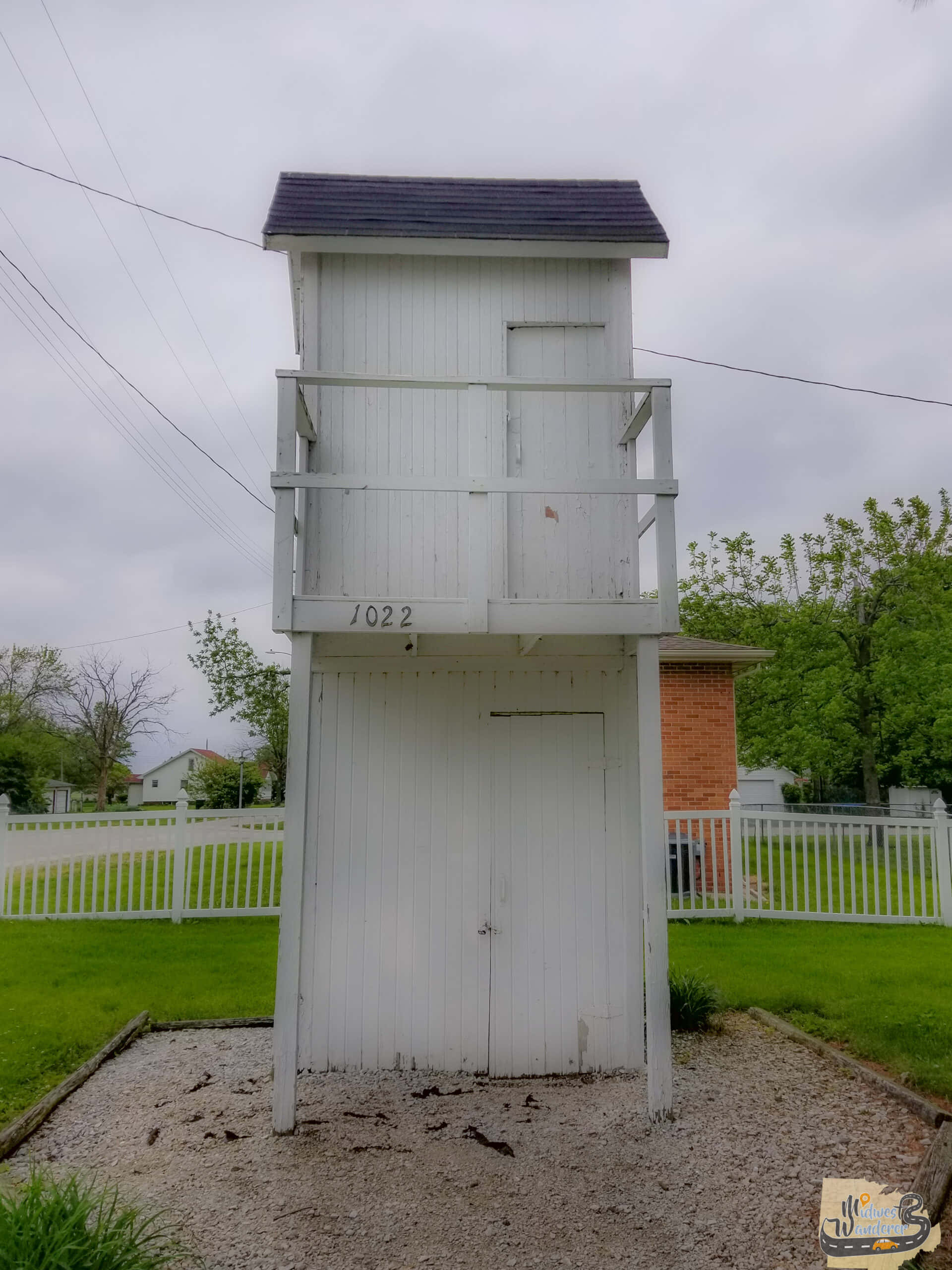 Outhouse 桌布 壁紙 — 一座高大的白色木製水塔，帶有深色屋頂，矗立在白色圍欄環繞的草地庭院中，天空多雲。