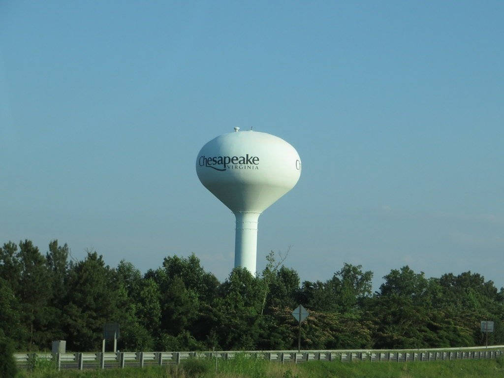 Virginia 桌布 壁紙 — 一座高大的白色水塔，上面標有Chesapeake Virginia，在晴朗的藍天下聳立在茂密的綠色樹線之上。