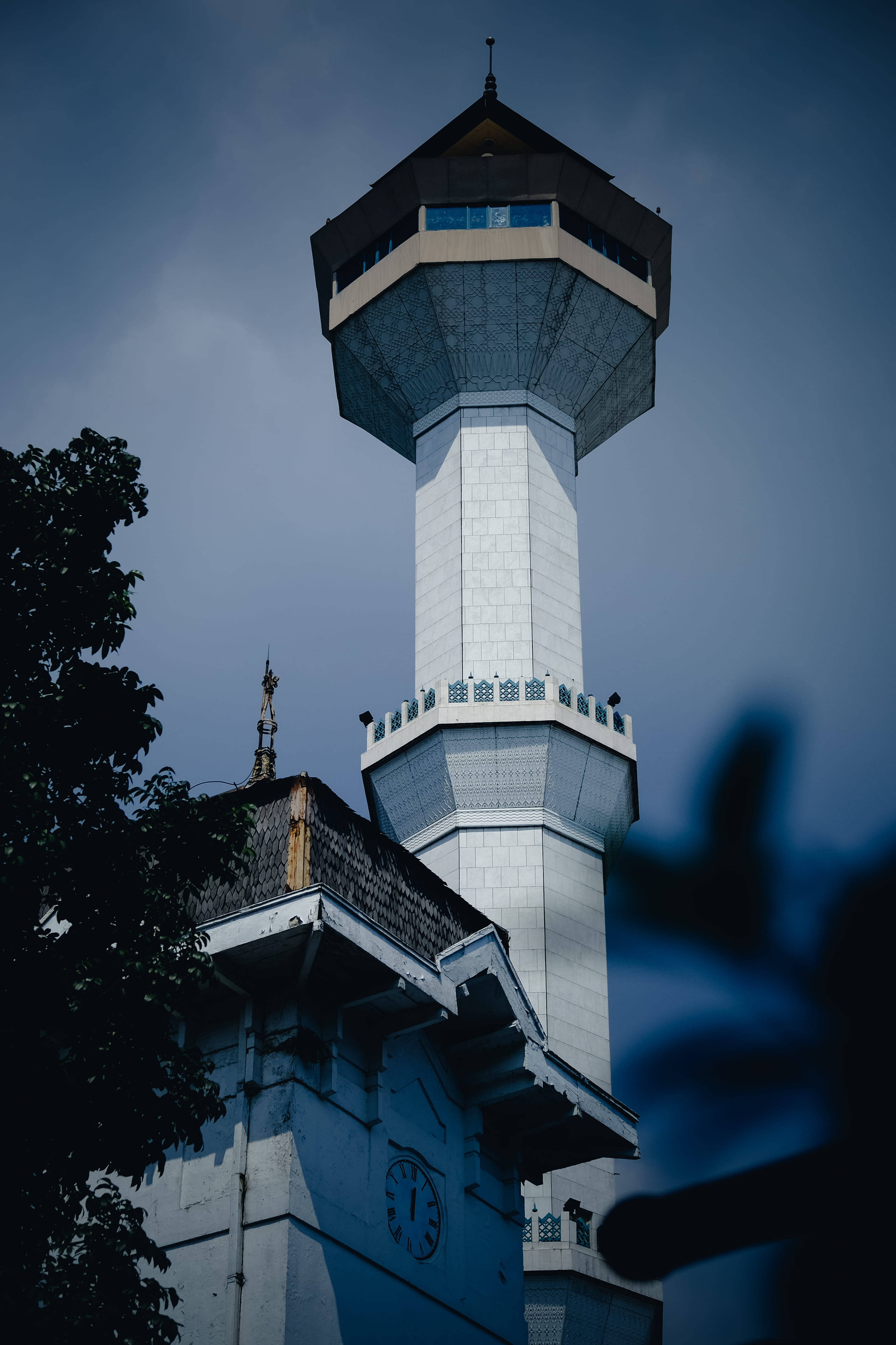 bandung 桌布 壁紙 — 一座高大的白色清真寺宣禮塔，帶有深色屋頂，在樹枝和模糊前景的映襯下，聳立在憂鬱的藍天中。