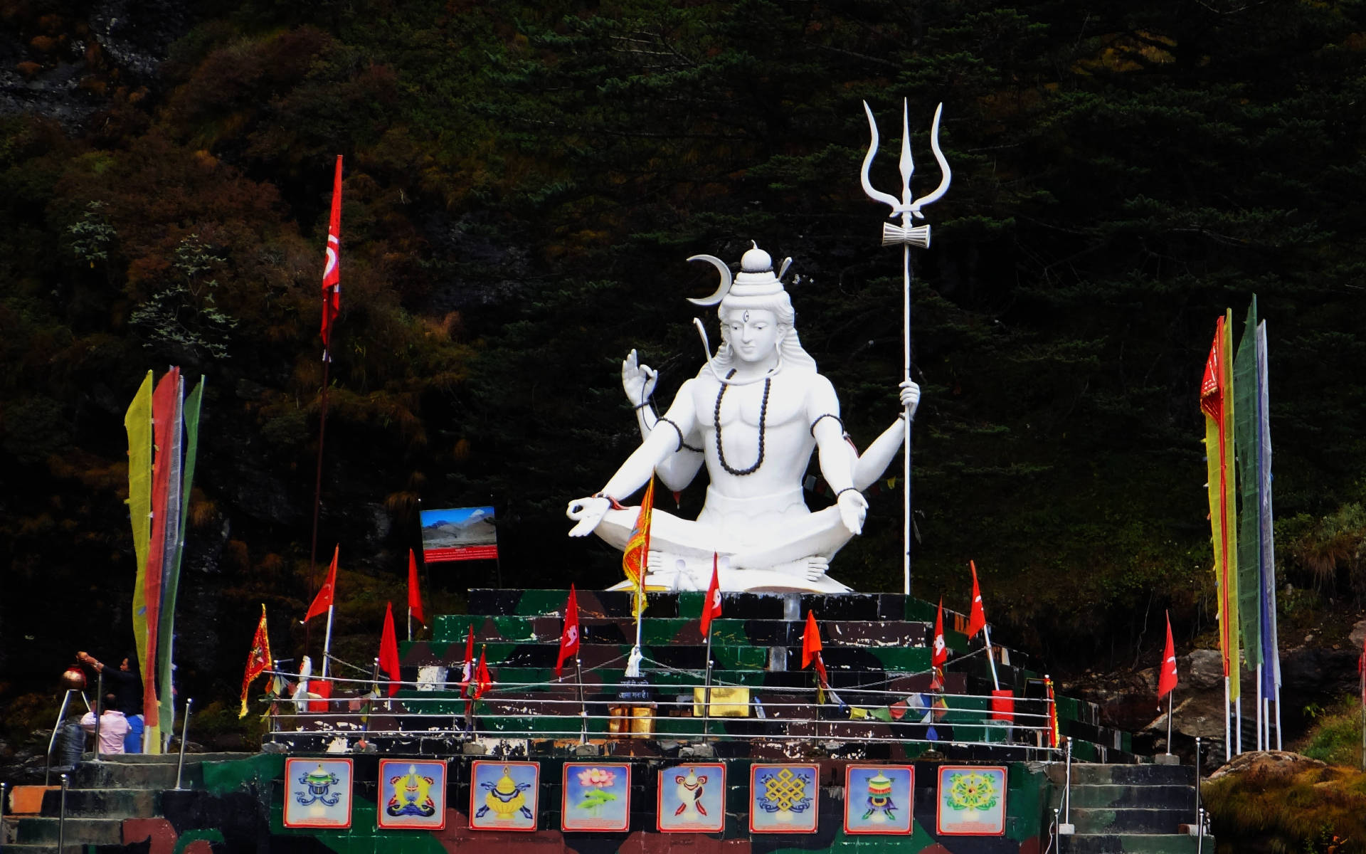 Indian 桌布 壁紙 — 一座高大的白色濕婆神像在裝飾平台上靜坐冥想，周圍環繞著色彩繽紛的經幡和茂密的森林。