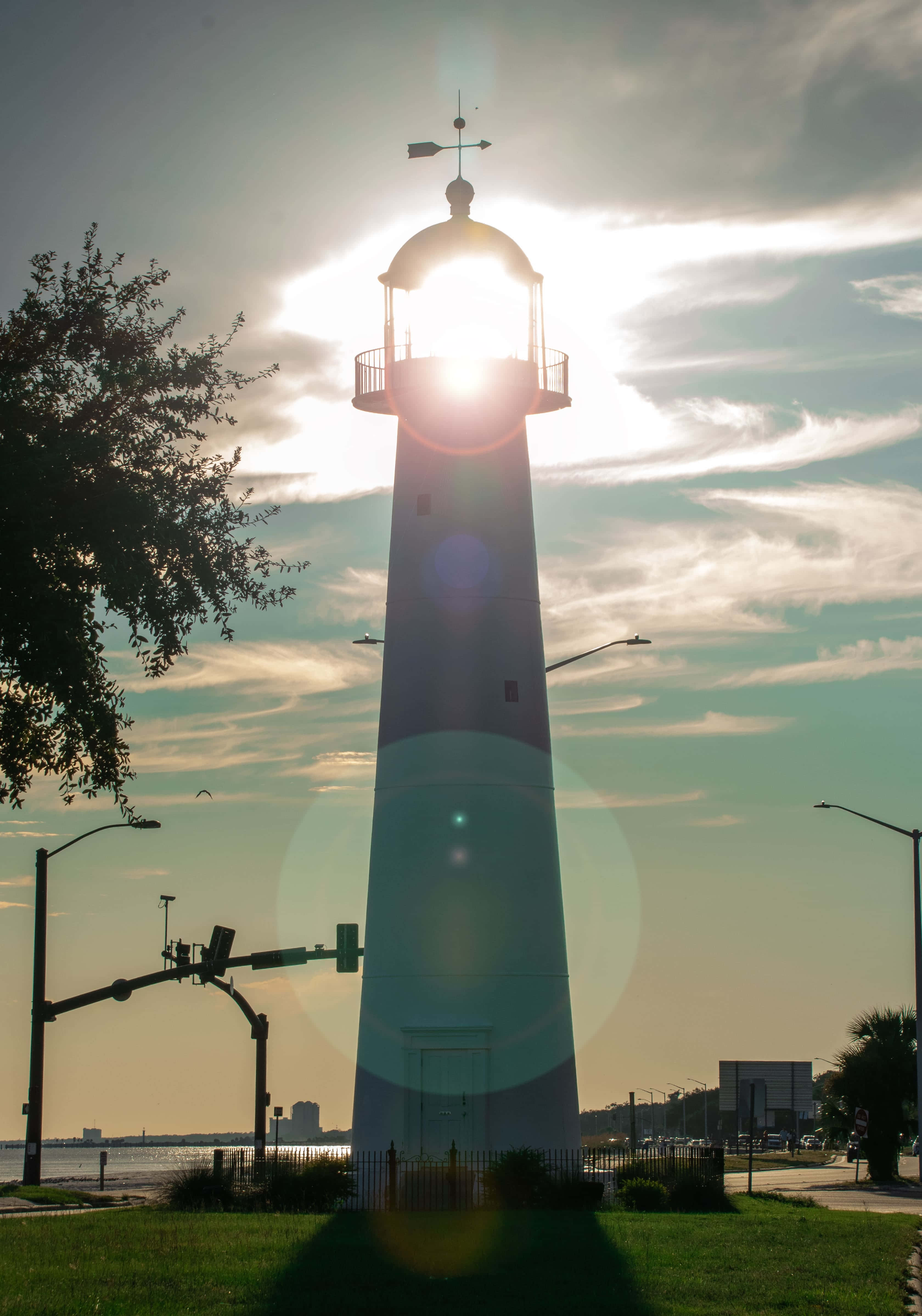 Lighthouse 桌布 壁紙 — 一座高大的白色燈塔在明亮的陽光爆發和鏡頭光暈中呈現剪影，在草地和附近的路燈上投下長長的影子。