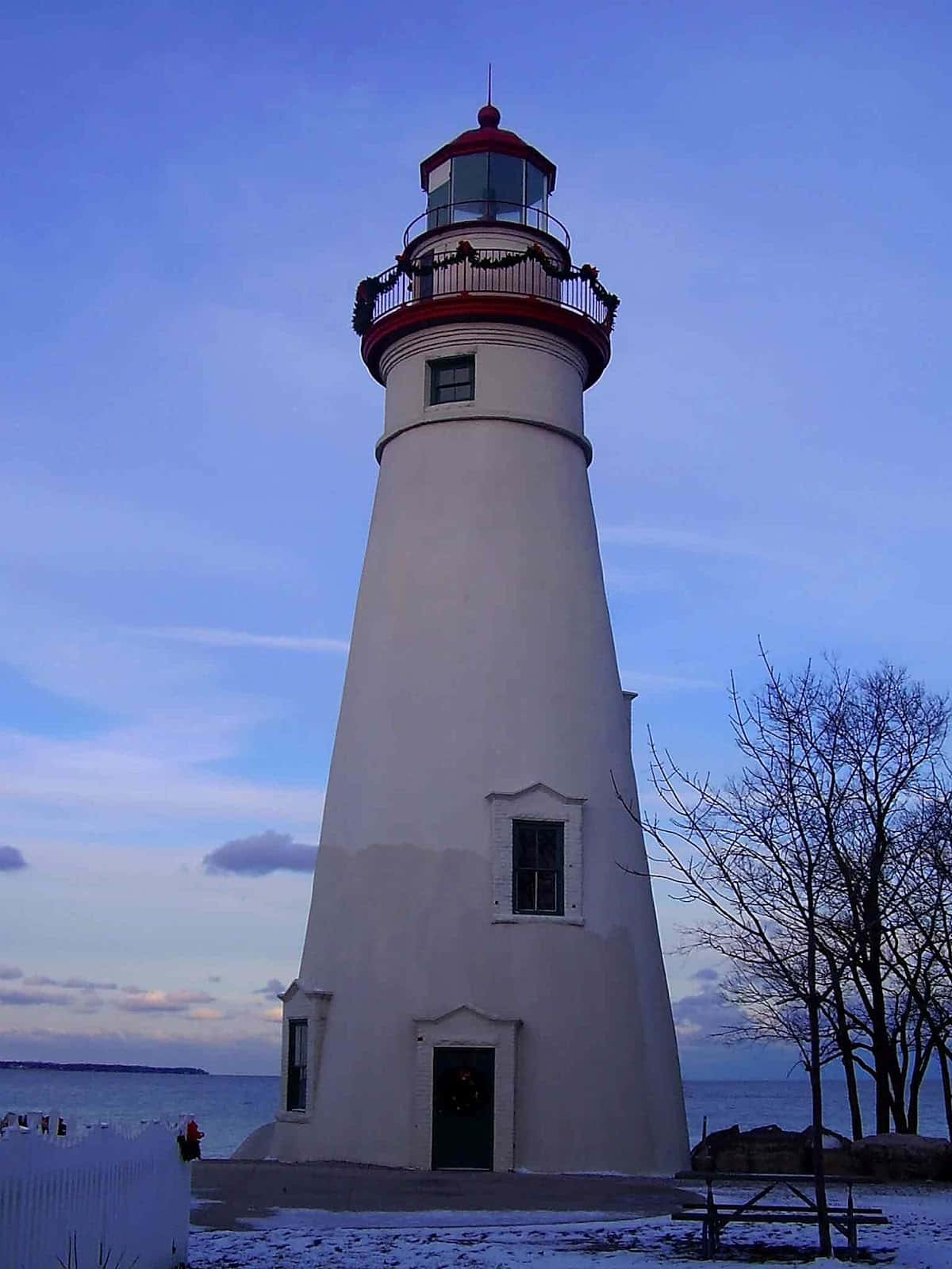Lighthouse 桌布 壁紙 — 一座高大的白色燈塔帶有紅色飾邊，矗立在暮色天空下，白雪覆蓋地面，光禿的樹木框住了場景。